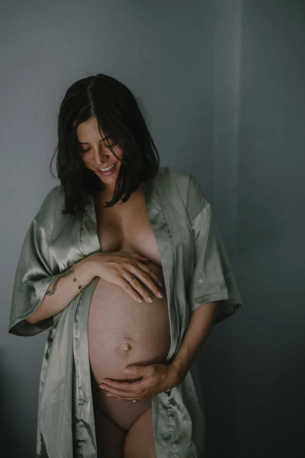 Schwangere Frau in Seidenk Kimono lächelnd, hält Bauch, steht vor grauer Wand.