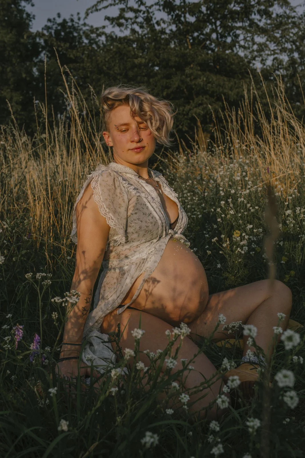 Schwangere junge Frau sitzt im Feld, umgeben von Blumen und Gräsern, bei Sonnenuntergang, trägt ein offenes Kleid, das ihren Bauch offen zeigt.