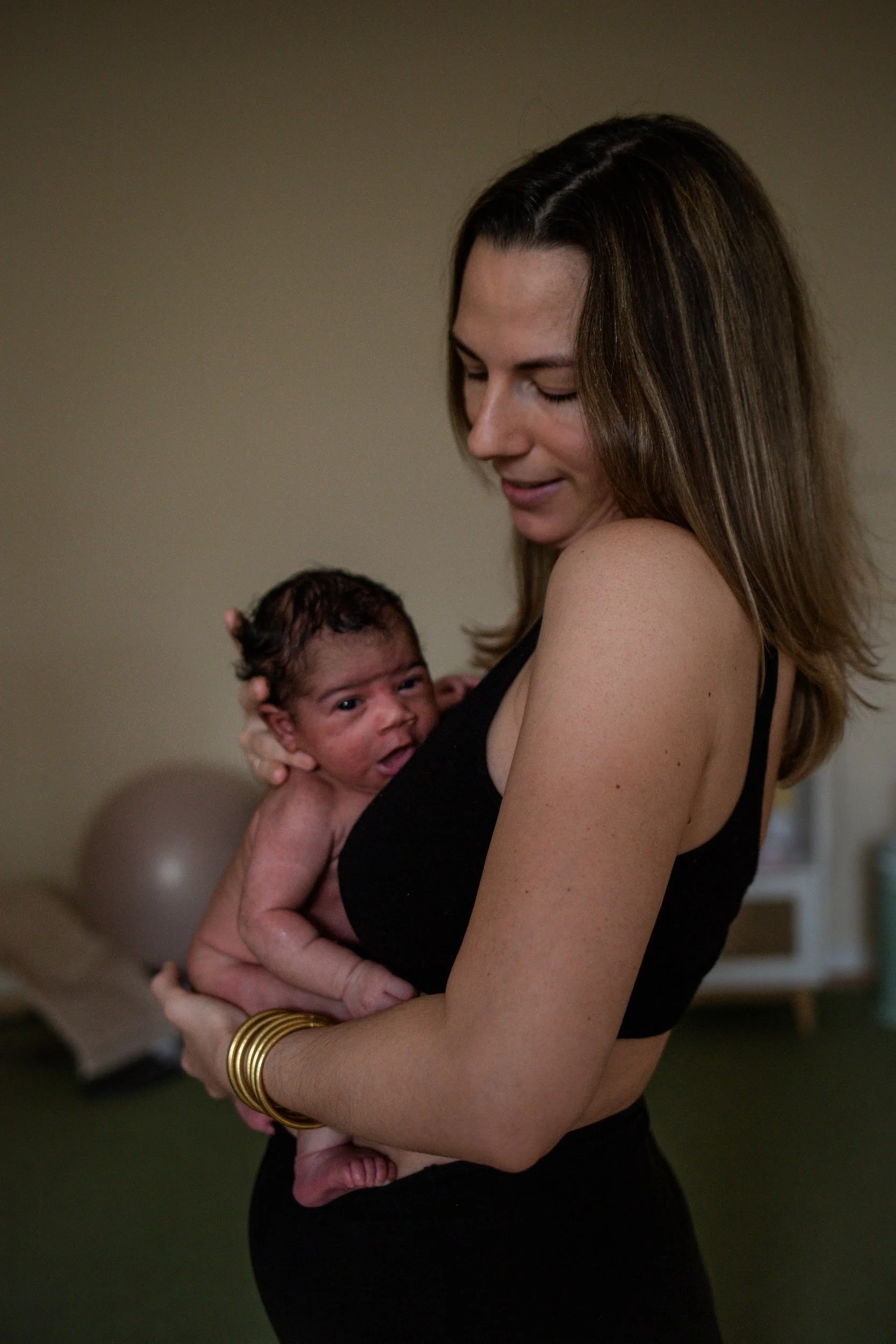 Eine Frau hält ein neugeborenes Baby an ihrer Brust, beide schauen nach oben, im Hintergrund ein Ball und ein Raum