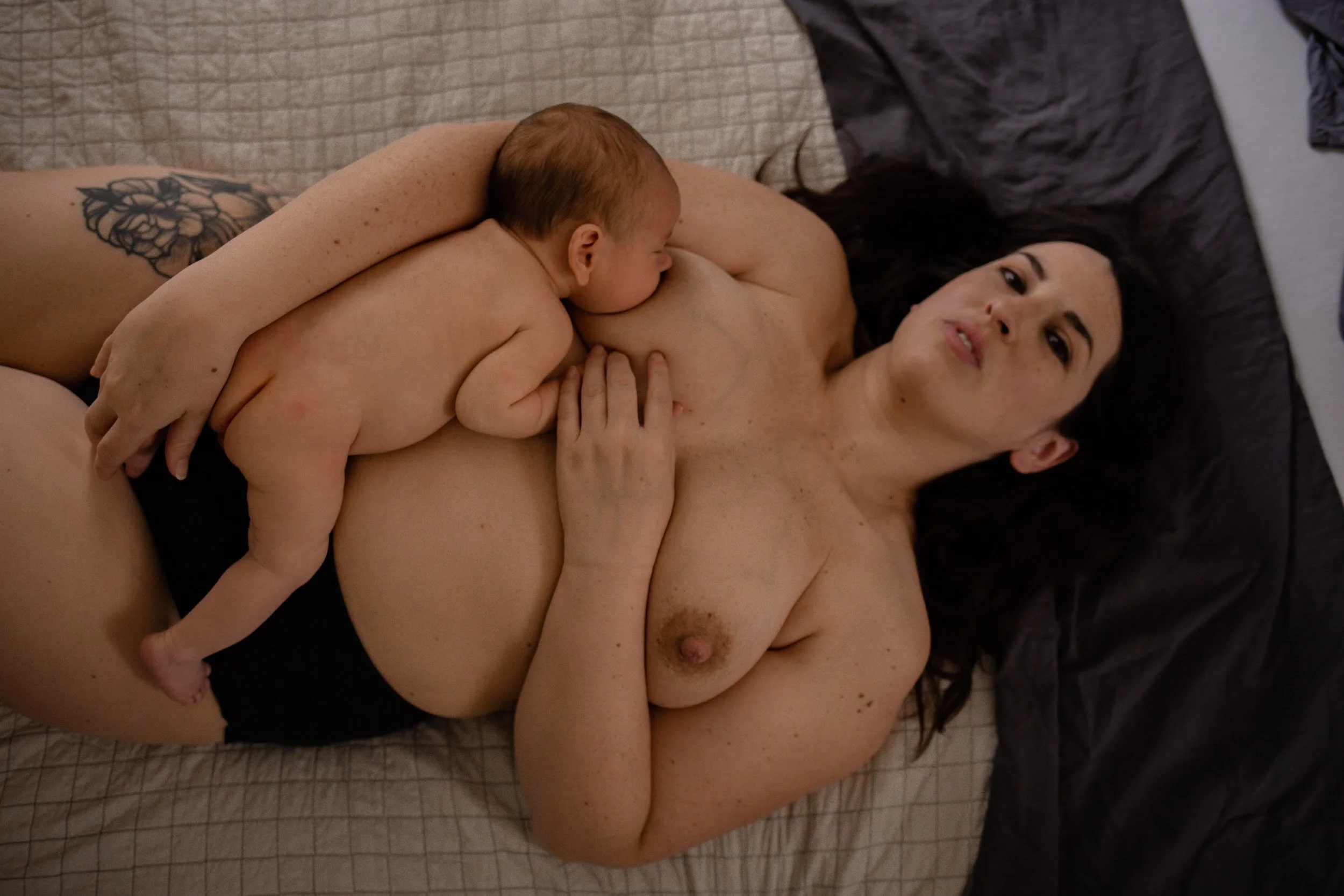 Eine nackte Frau liegt auf einem Bett und isst von einem Baby, das auf ihrer Brust liegt. Das Baby ist nackt und der Frau sind dunkle Haare und Hautflecken sichtbar.