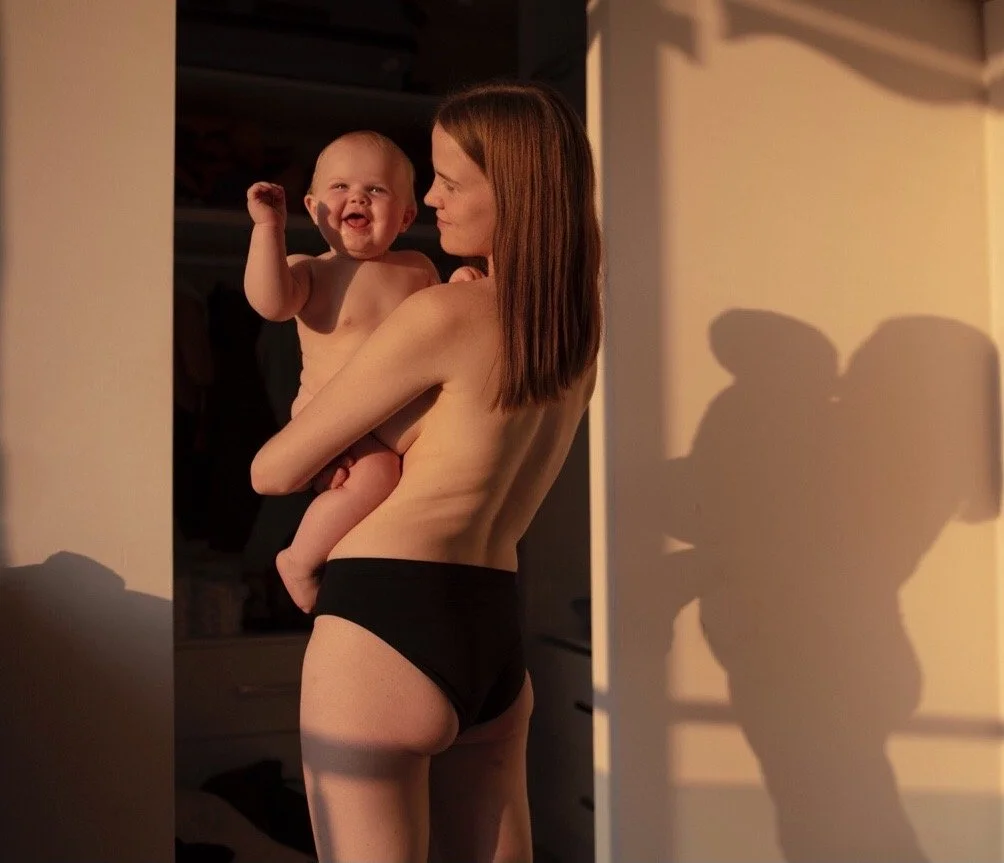 Eine Frau mit roten Haaren hält ein lachendes, nacktes Baby im Arm vor einer hellen Wand, beide werfen Schatten an die Wand, bei Sonnenlicht.