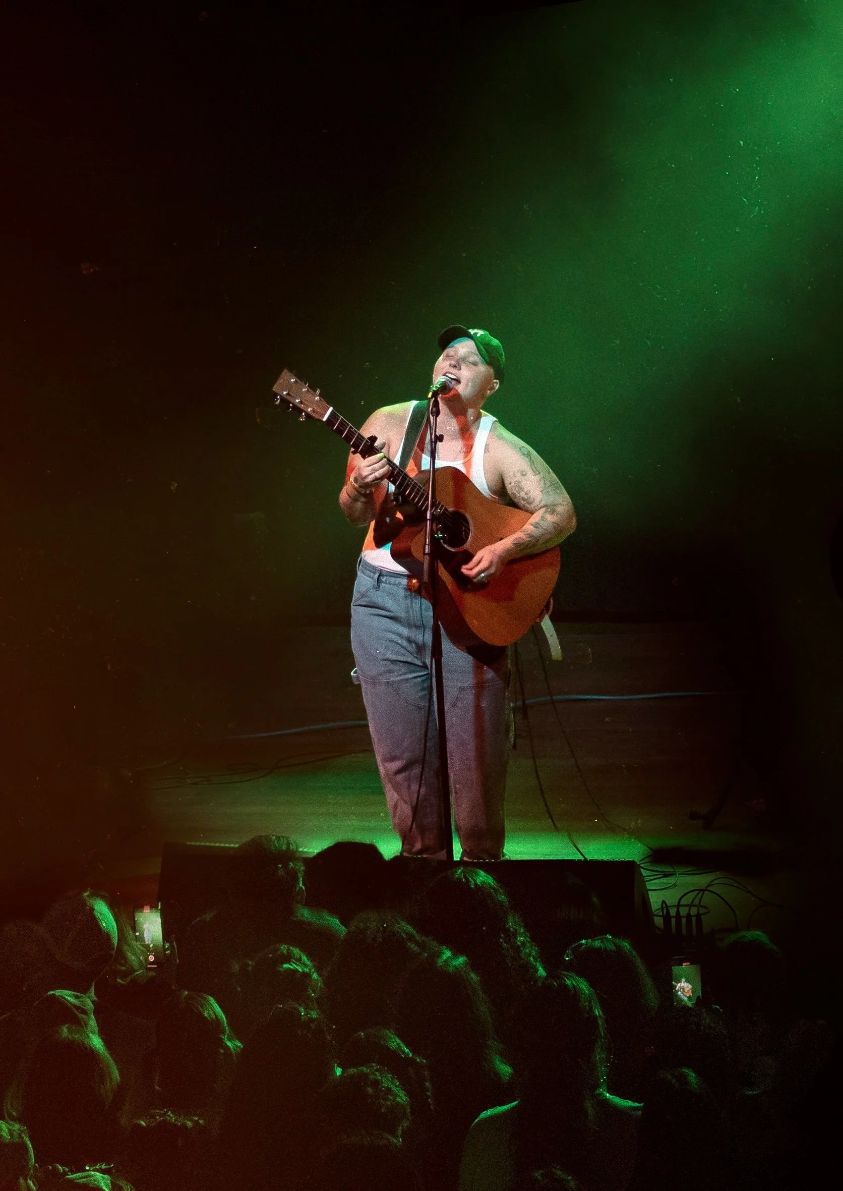 Famous queer music artist miki ratsula singing into a microphone and playing an acoustic guitar on stage, with an audience in front. The stage is lit with green lighting.