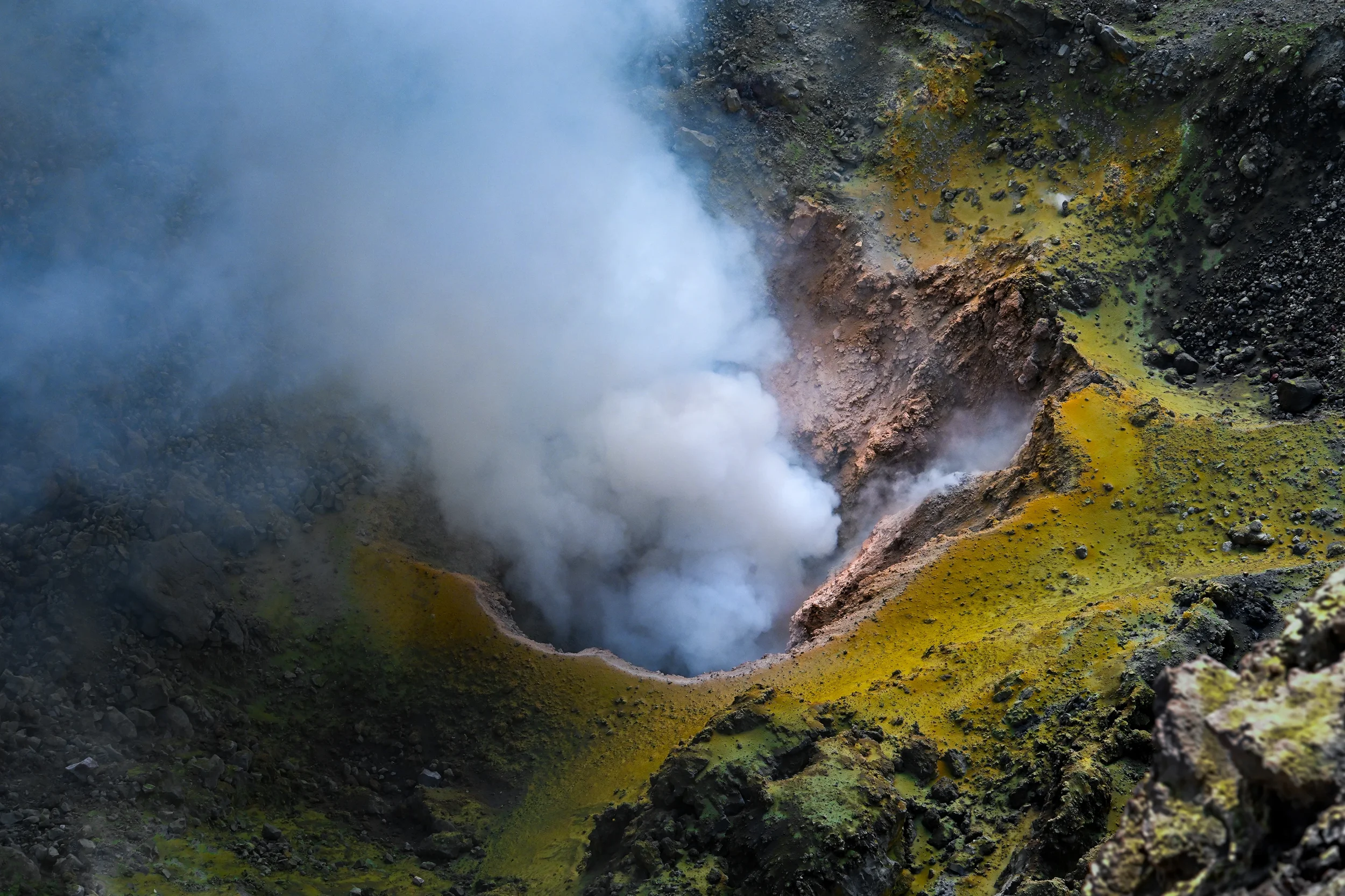 A volcanic crater emitting steam and gas, with surrounding rocky, green and yellow sulfur-covered terrain, known as mount etna in sicily, italy.
