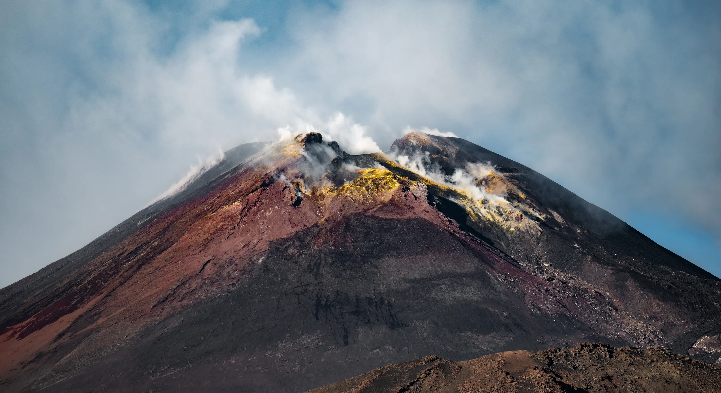 The top of a volcano showing yellow, sulfur-covered terrain, red and purple volcanic rock, and steam rising from the summit crater of mount etna in sicily, italy.