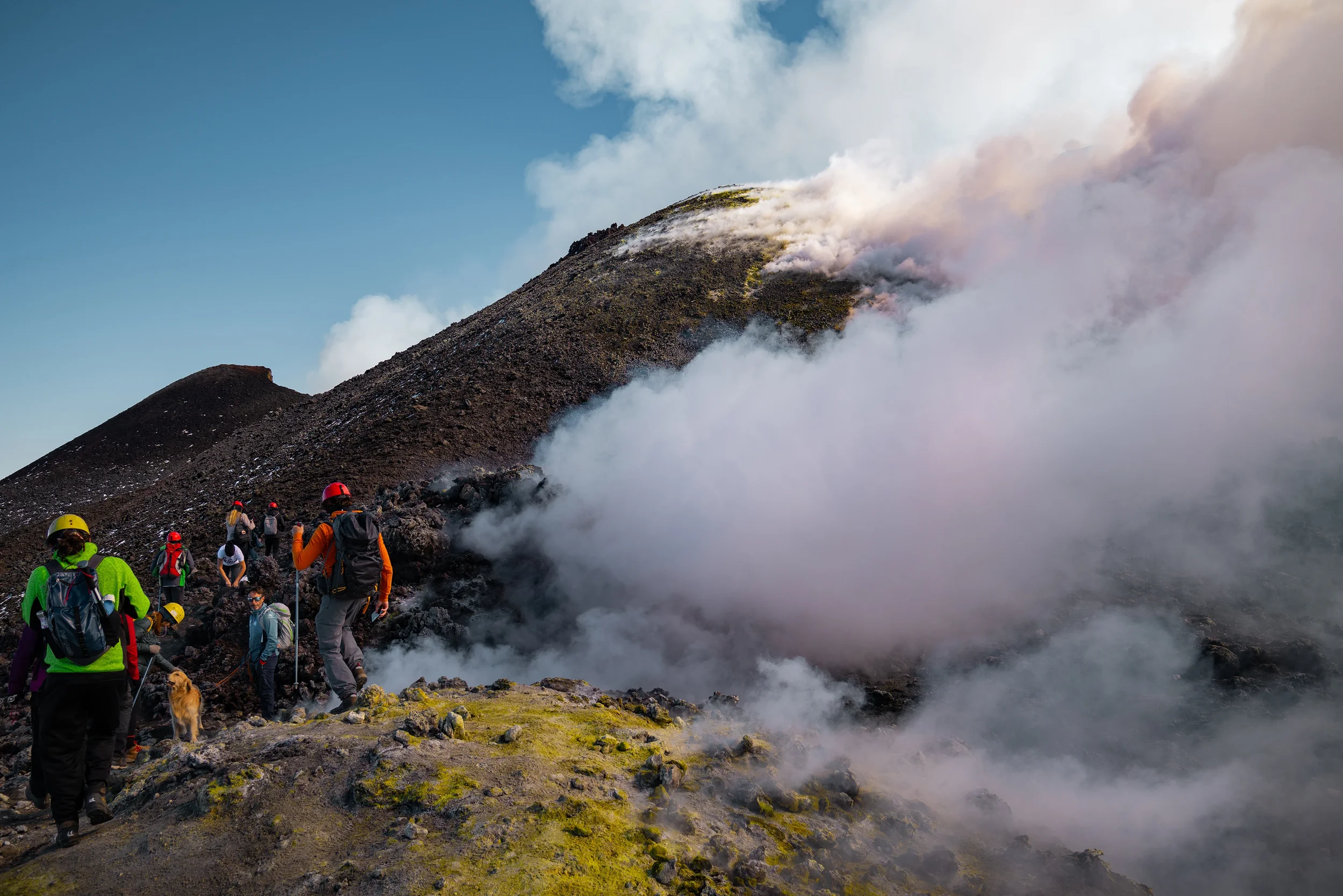 Group of hikers and a dog ascending a volcanic mountain, mount etna, with sulfuric steam and dark rocky terrain in sicily, italy.