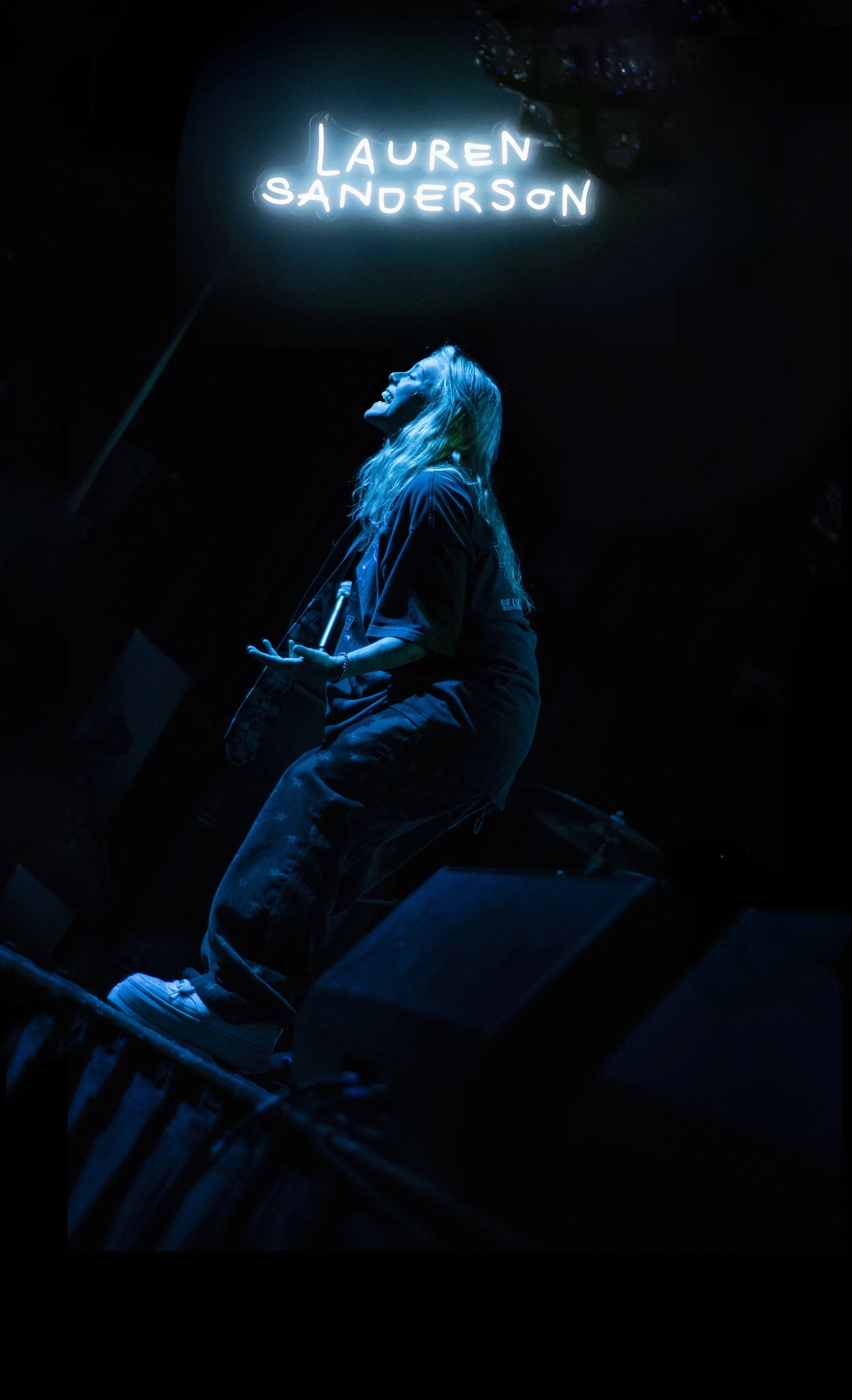 A woman with long blonde hair performing on stage, illuminated by blue light, with a neon sign above her that reads 'LAUREN SANDERSON'.