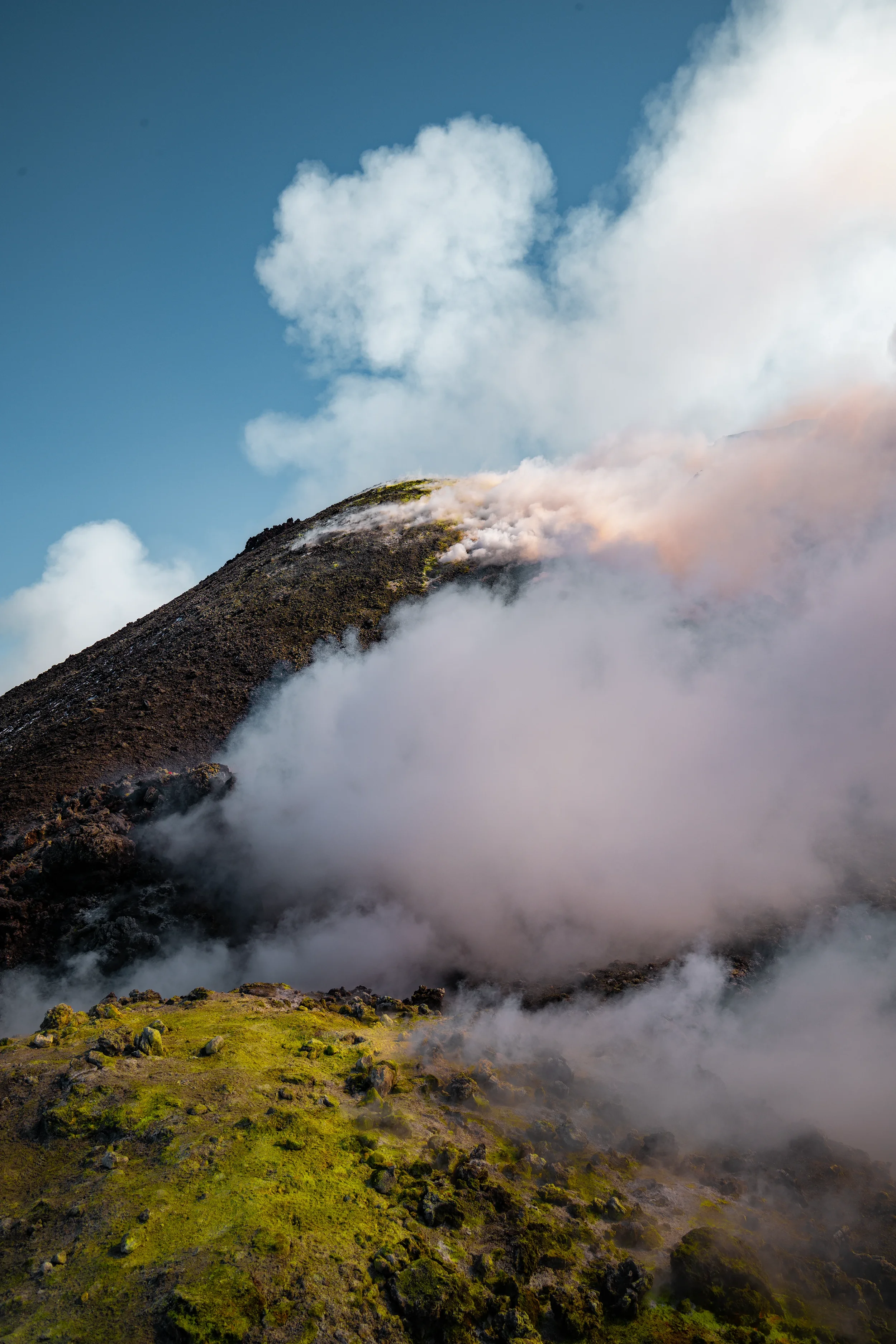 Mount etna volcano in sicily, italy, erupting with smoke and ash, surrounded by clouds and mist, with green mossy ground at the base.