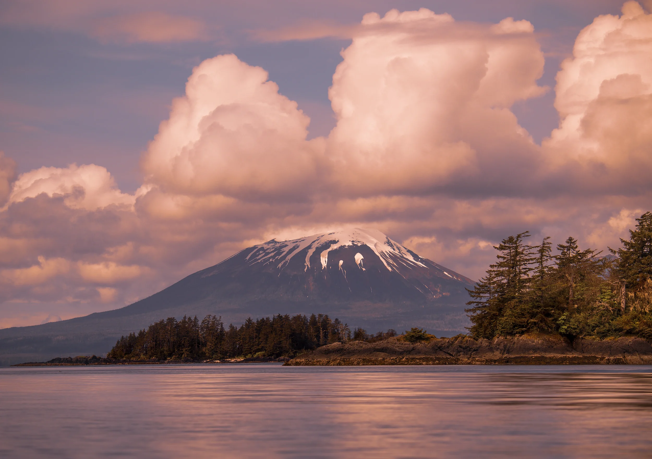 A snow-capped volcanic mountain with clouds above, green trees on the rocky shoreline, and calm water in the foreground, also known as mount edgecumbe, located in sitka, alaska.