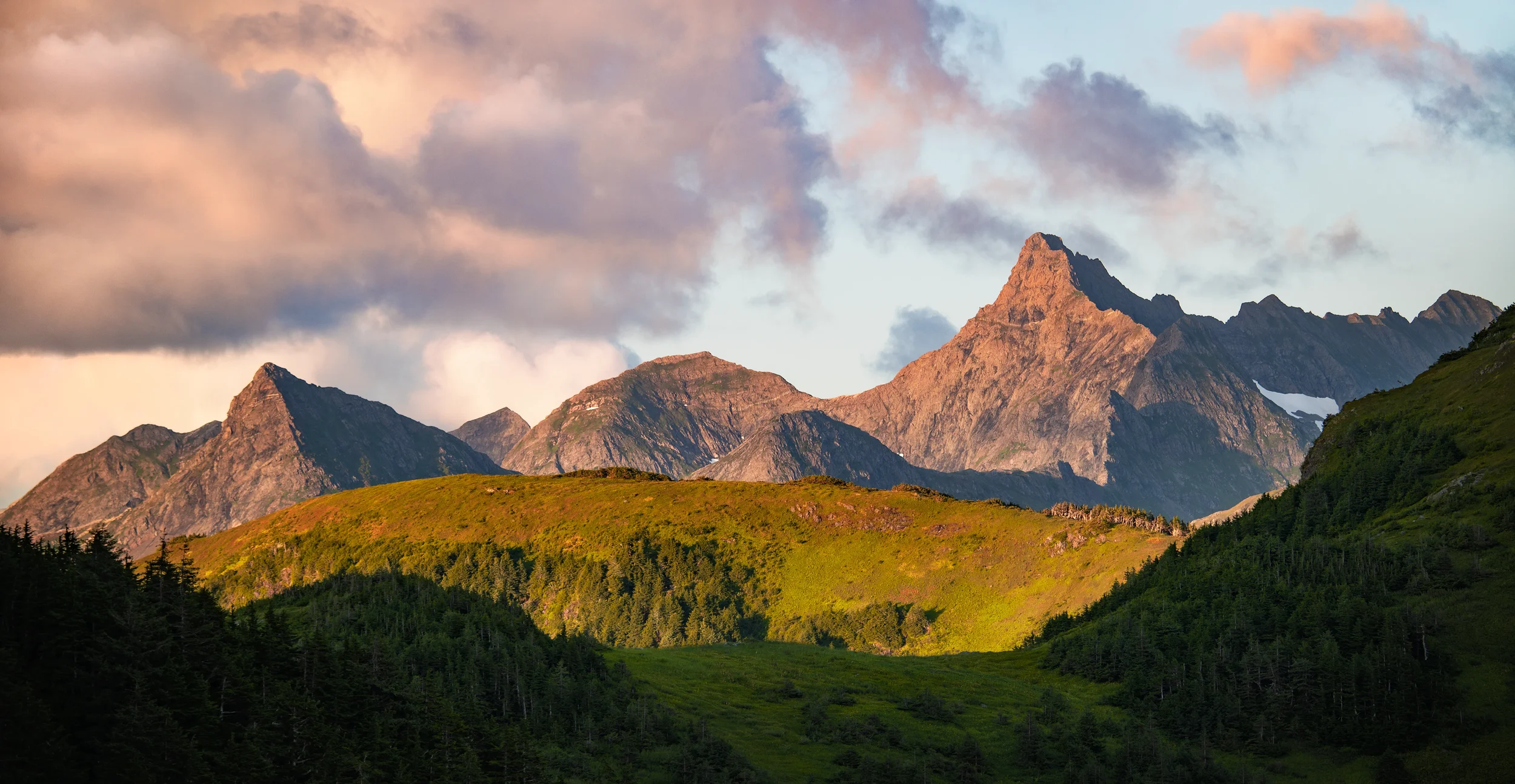 Sunset over rugged mountains and green hills, with partly cloudy sky.