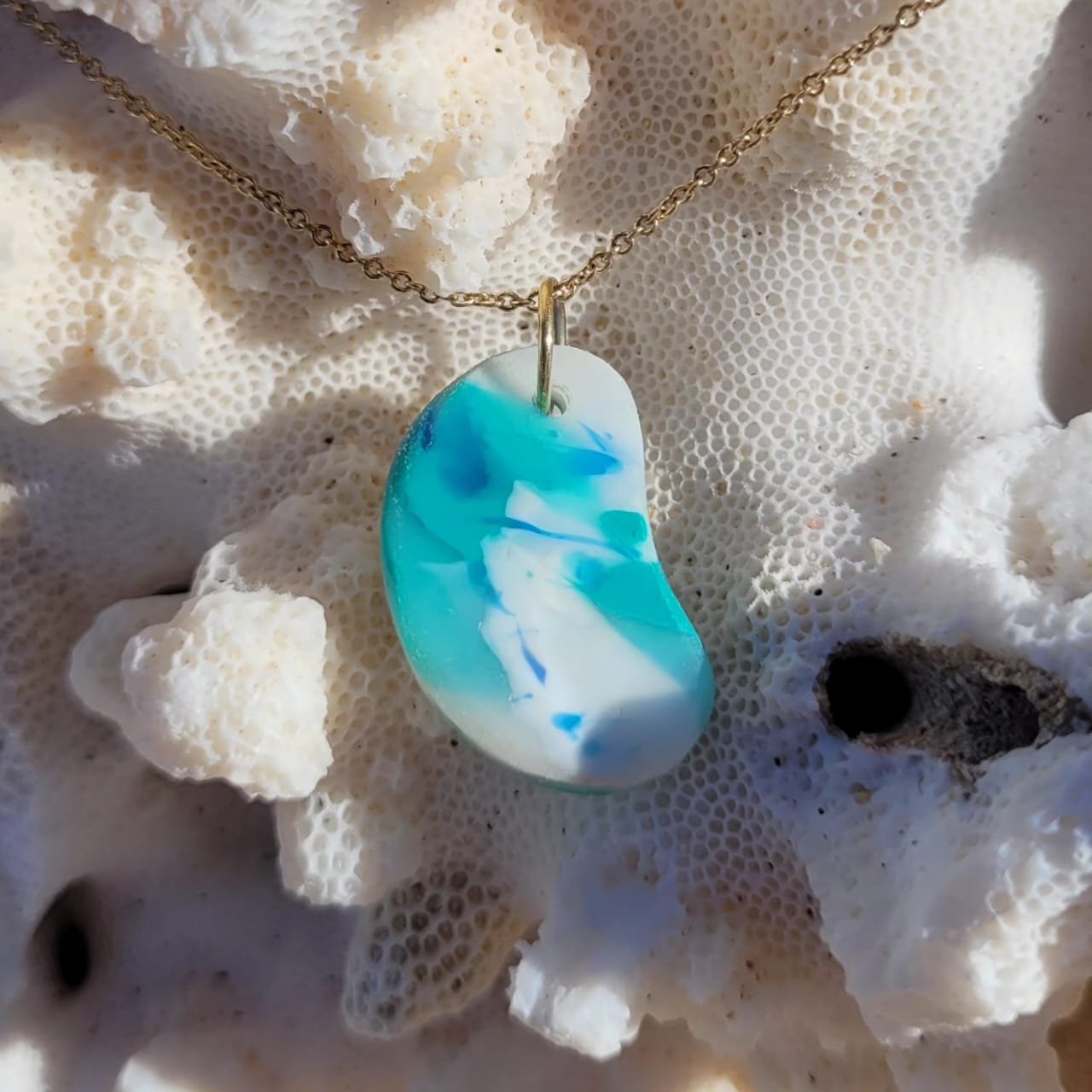 Collar de cadena dorada con un dije de plástico azul y blanca, sobre coral blanco y algunas rocas negras.