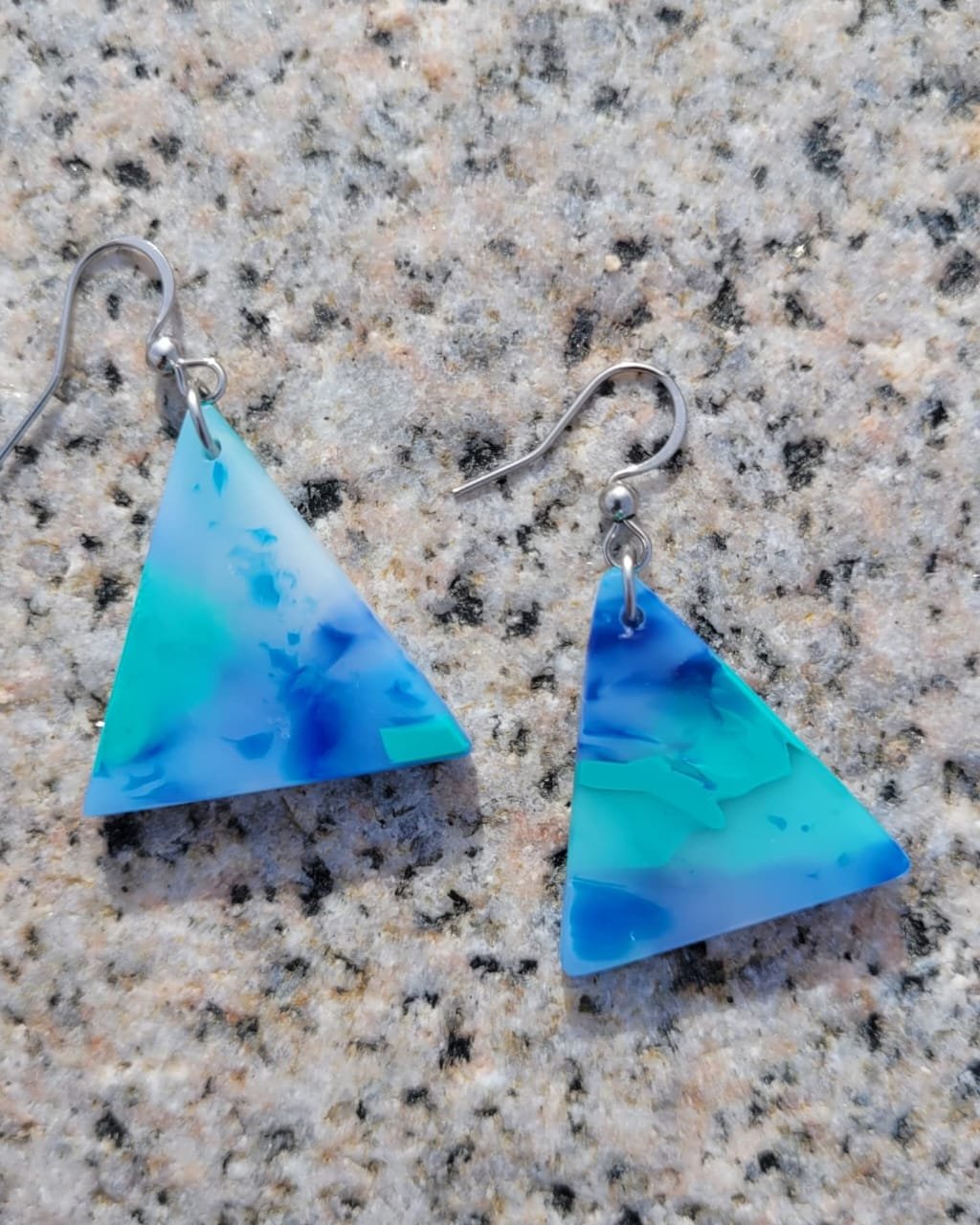 Aros con aretes de acrílico en forma de triángulo de color azul con detalles en tonos blancos y oscuros, colocados sobre una superficie de granito.