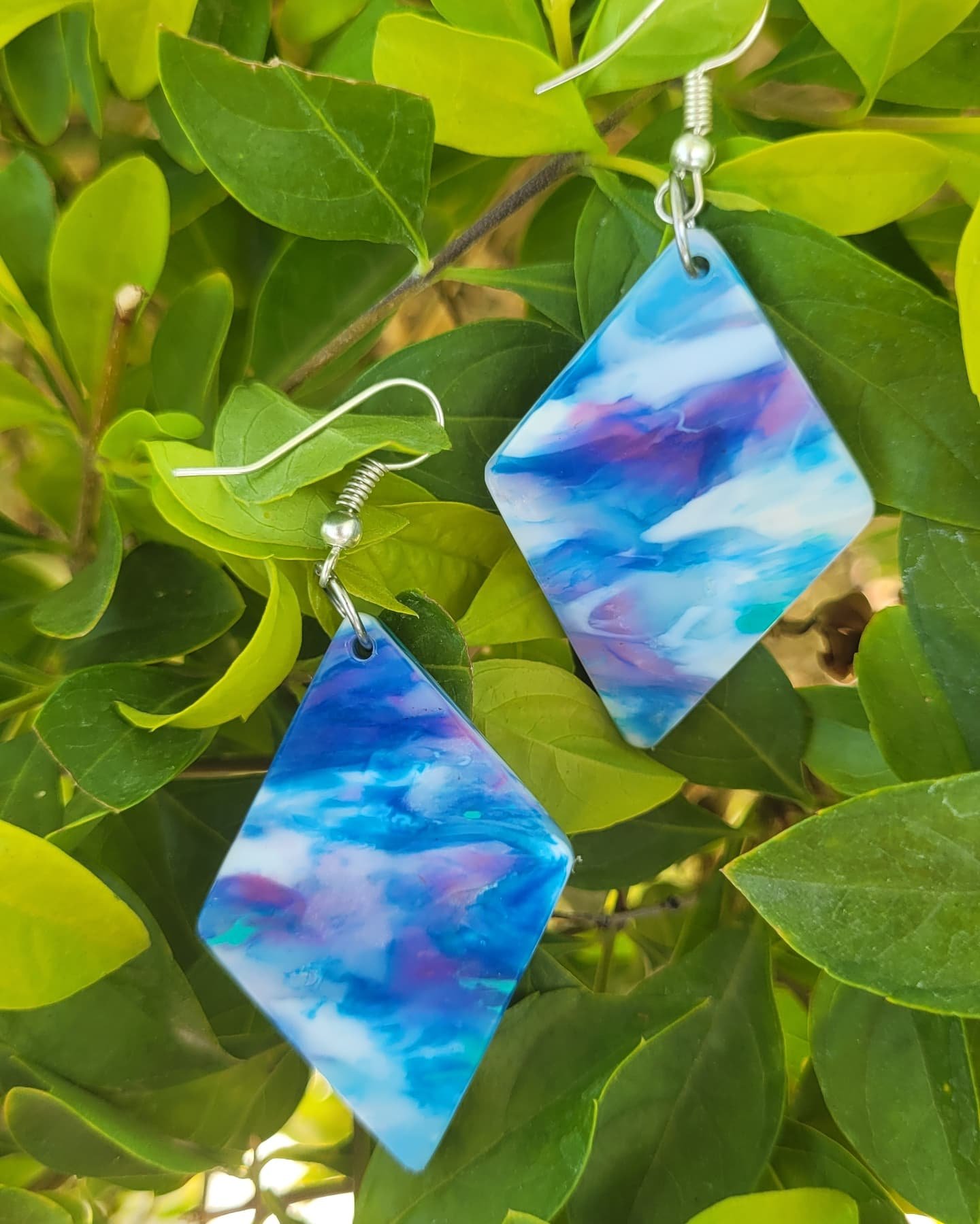 Aretes de color azul con diseño de nubes, colgados en una planta con hojas verdes.