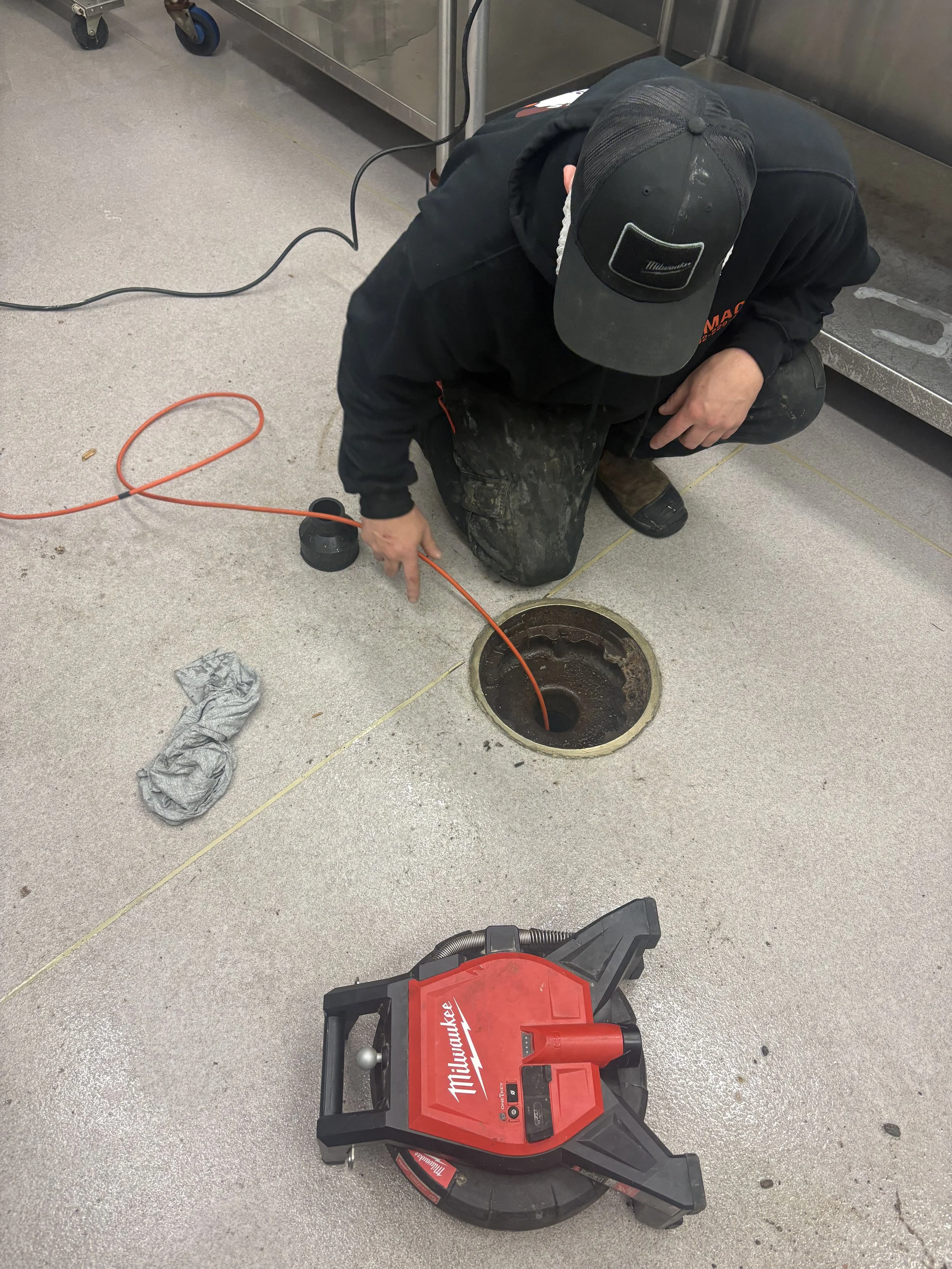 A worker kneeling on a concrete floor, using a specialized vacuum tool to clear debris from a sewer drain opening. The worker is wearing a black hoodie and cap, with a red Milwaukee vacuum machine nearby, and various cords on the floor.