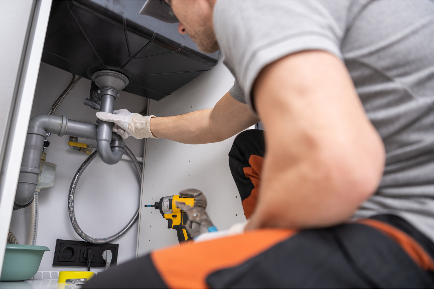 A plumber fixing pipes under a kitchen sink using a cordless drill.
