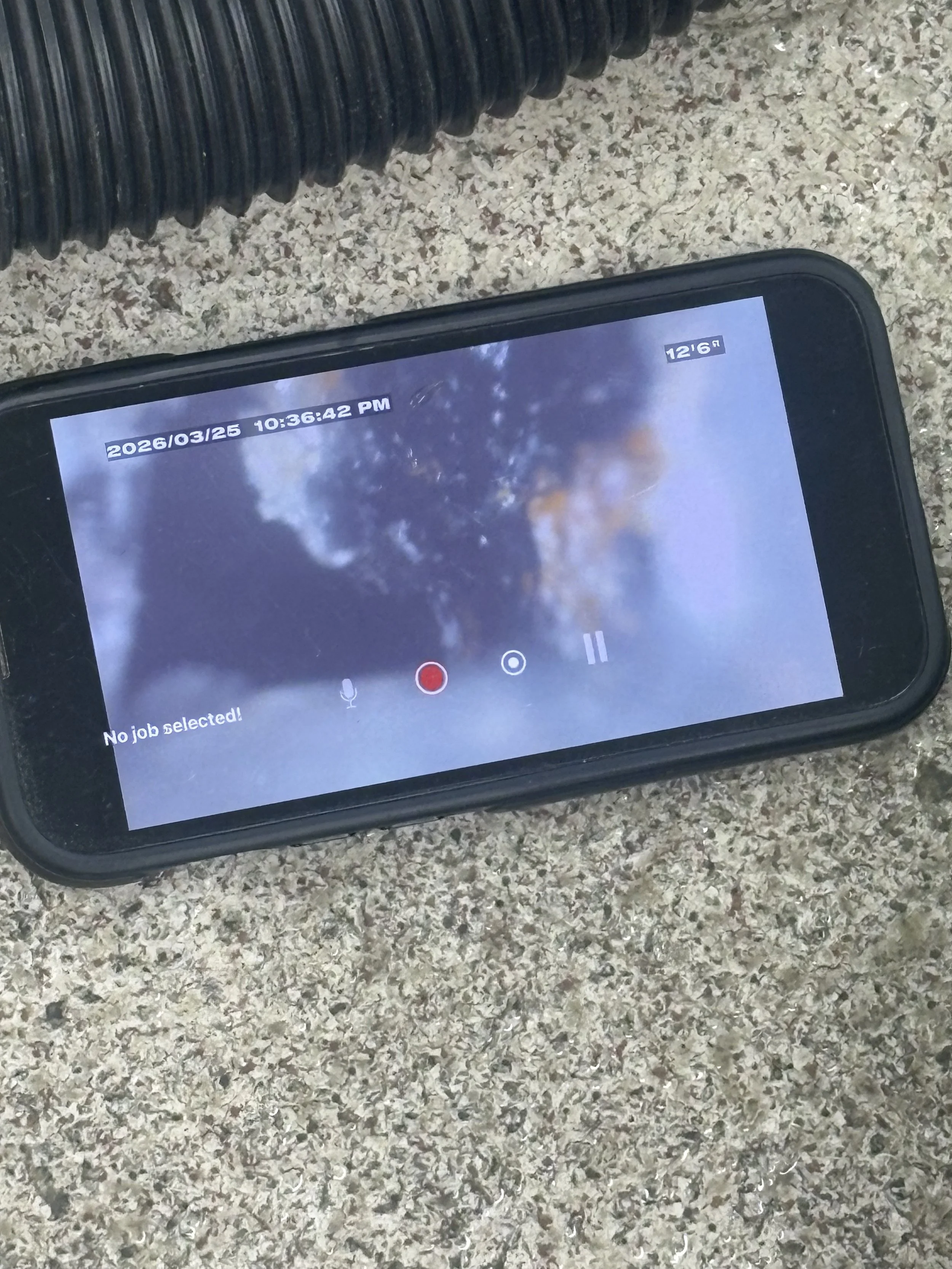 A smartphone lying on textured ground displaying a paused video or recording screen, with a blurred background and timestamp, showing no selected job.
