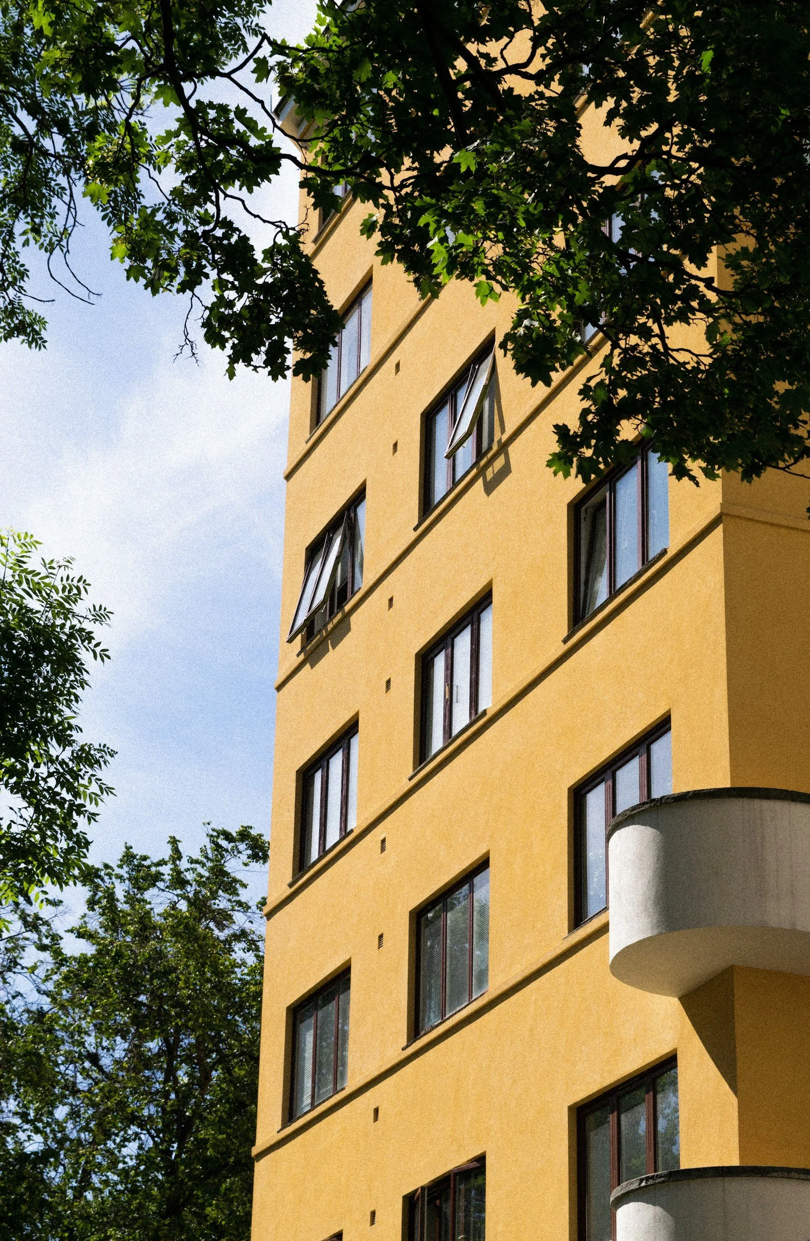 Mehrstöckiges gelbes Wohngebäude mit mehreren offenen Fenstern, umgeben von grünen Bäumen unter einem blauen Himmel.