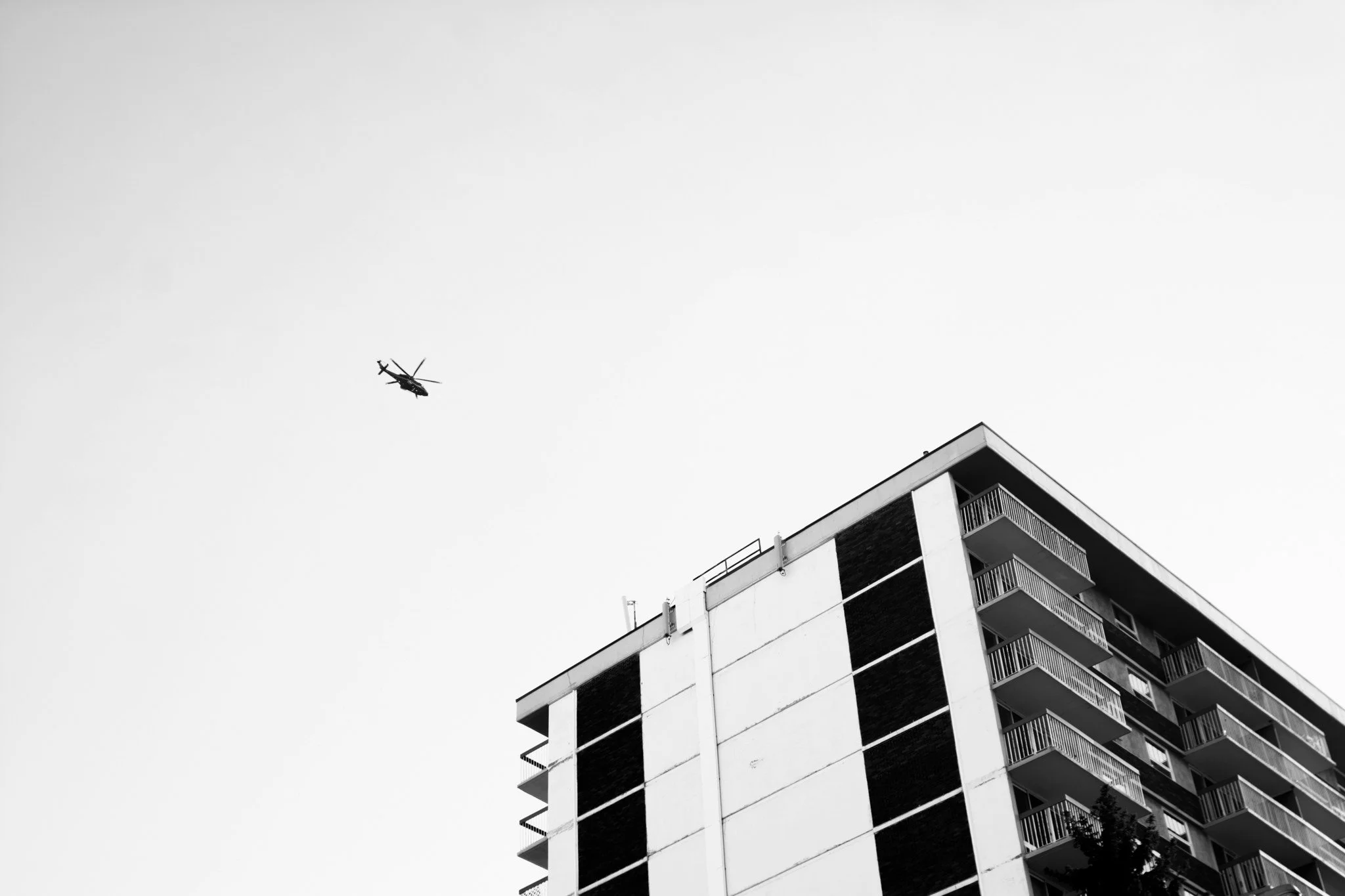 Ein schwarzer Hubschrauber fliegt über ein Mehrfamilienhaus mit Balkonen in Schwarz-Weiß