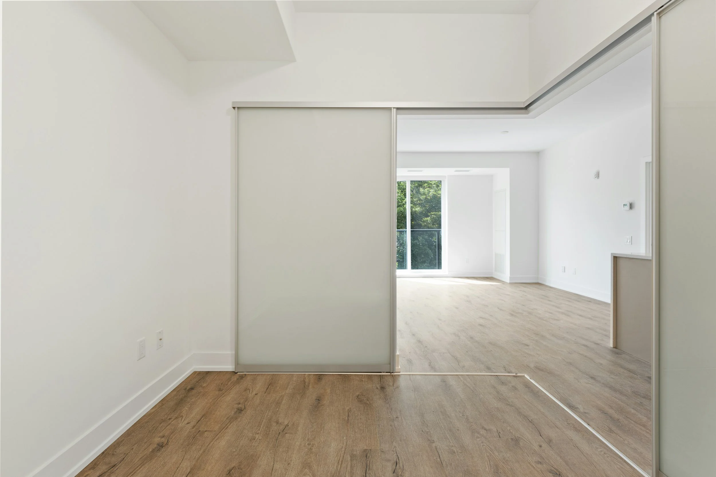 Ein helles, leeres Zimmer mit Holzboden und einer halb offenen Trennwand mit einer Glastür, die ins Wohnzimmer führt, mit großen Fenstern im Hintergrund, die viel Tageslicht hereinlassen.