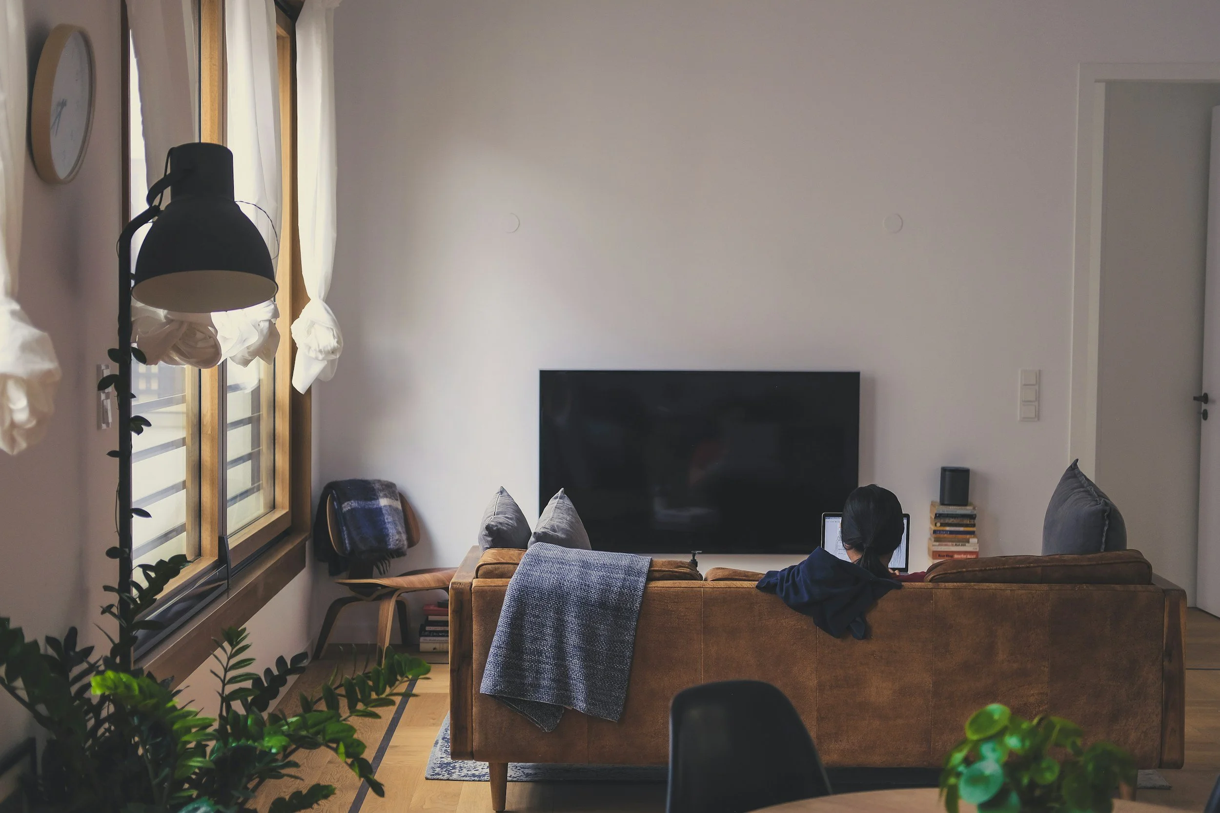 Wohnzimmer mit braunem Sofa, TV, Laptop, Bücher, Fenster mit weißen Vorhängen, Uhr an der Wand, grüne Pflanze im Vordergrund.