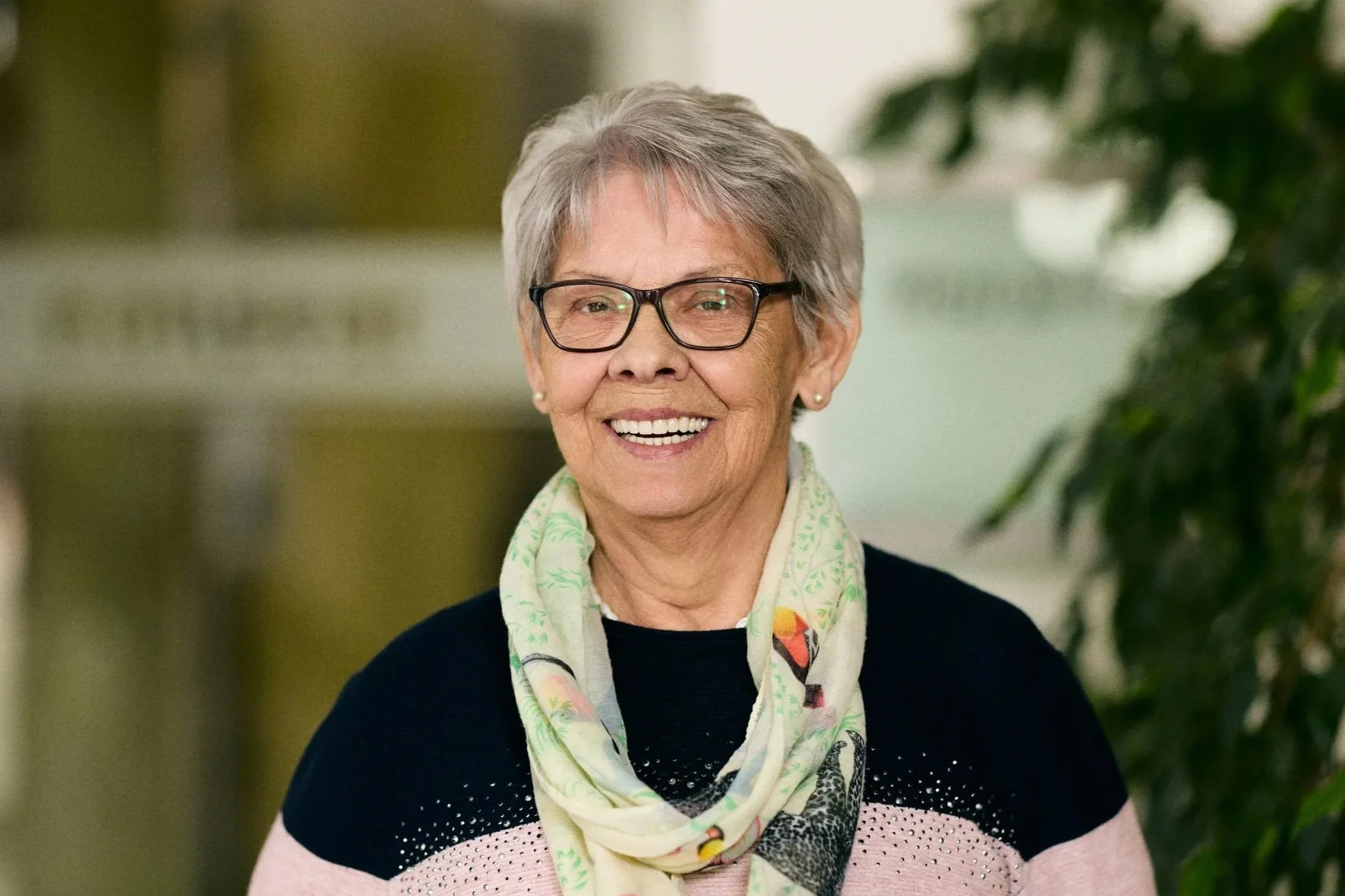 Eine Frau mit Brille und dunkelrotem Haar, die eine gelbe Kleid trägt und auf einer Brüstung lehnt, im Hintergrund Bäume.