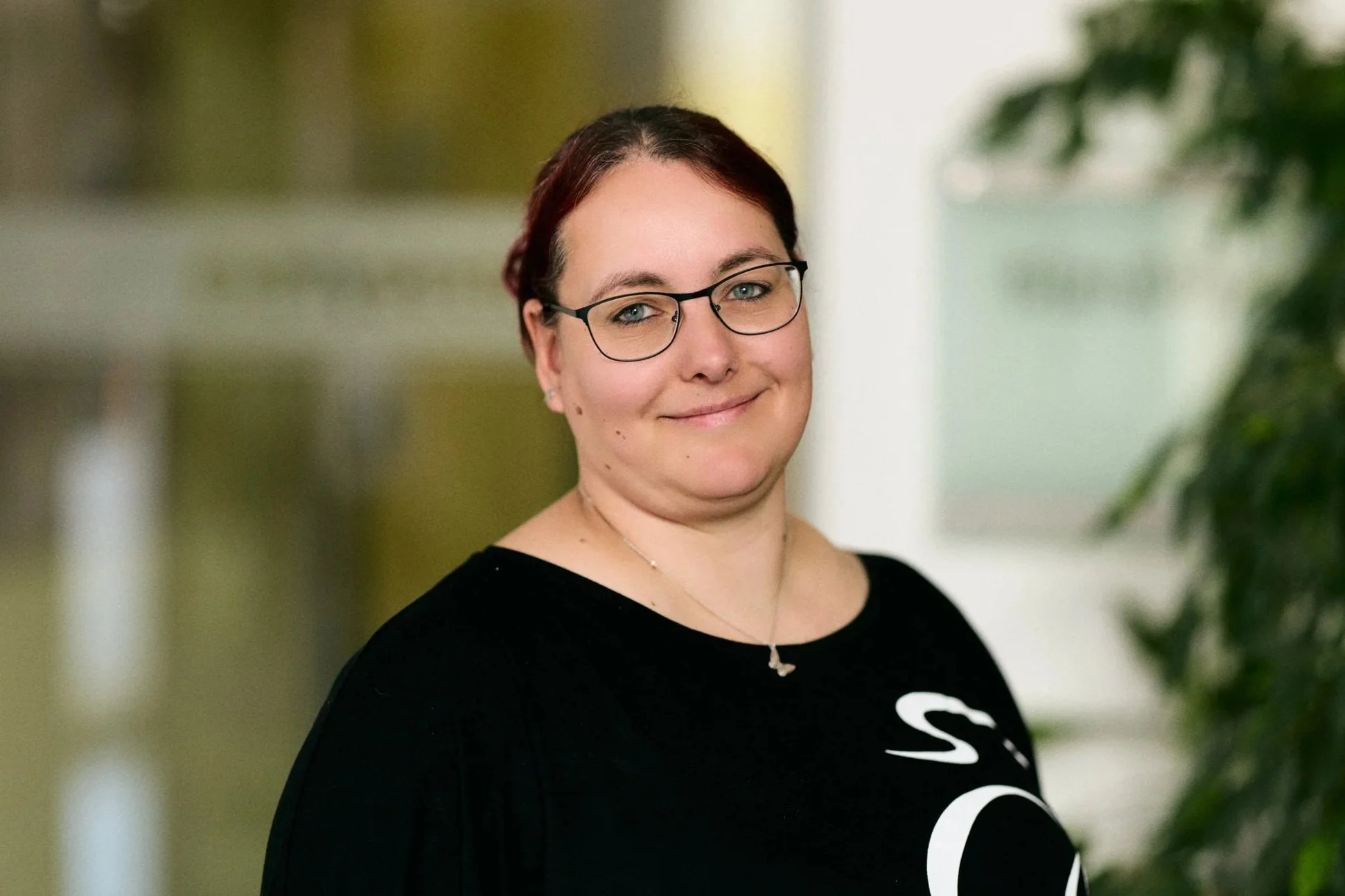 Porträt einer lächelnden Frau in einem hellen Büro, trägt eine schwarze Bluse, eine gestreifte Jacke und eine goldene Halskette.