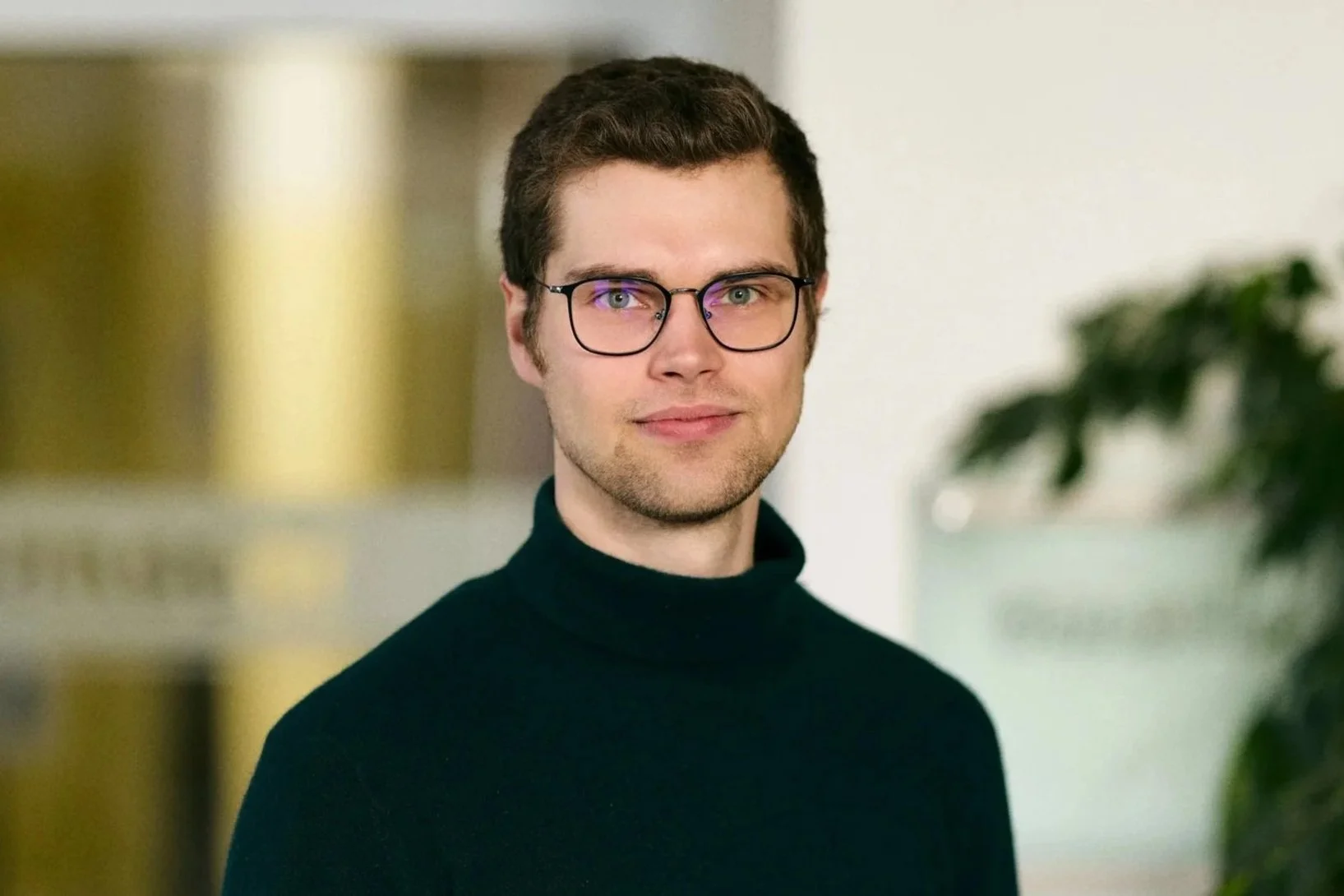 Ein junger Mann mit lockigem braunem Haar und einem leichten Lächeln, trägt einen schwarzen Pullover vor weißem Hintergrund.
