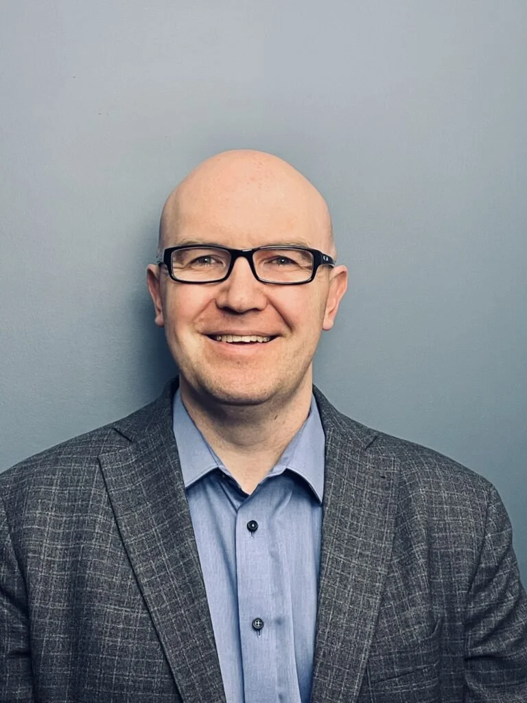 A smiling man wearing glasses and a checked blazer over a blue dress shirt, standing against a plain gray wall.