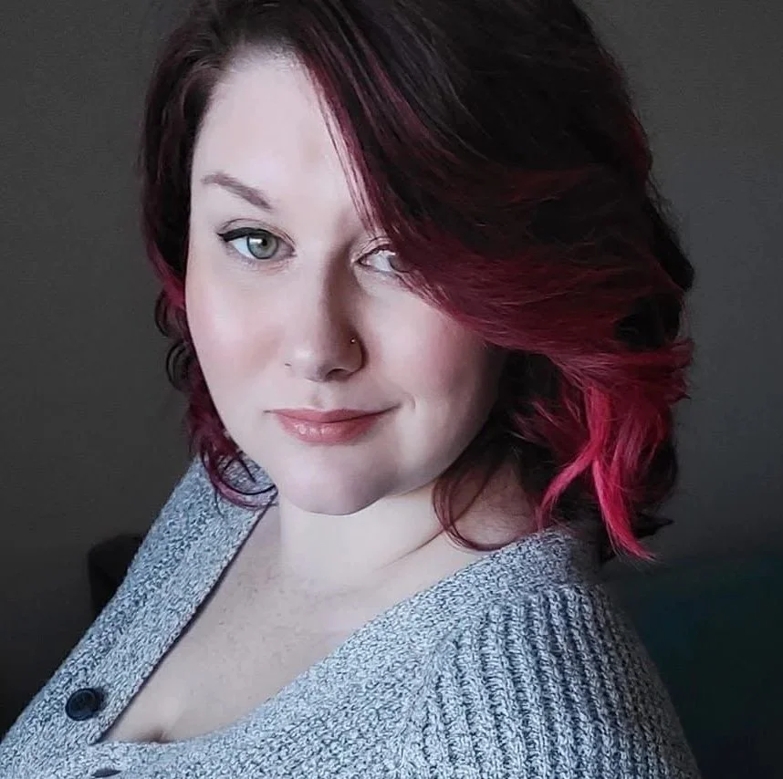 A woman with shoulder-length dark red hair with pink highlights, light skin, and blue eyes, wearing a gray knitted sweater, posing in front of a dark background.