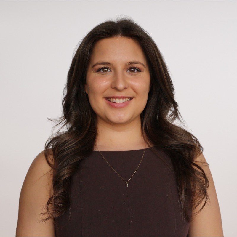 A young woman with long, dark brown hair styled in loose waves, smiling against a plain white background, wearing a sleeveless dark brown top and a delicate necklace.