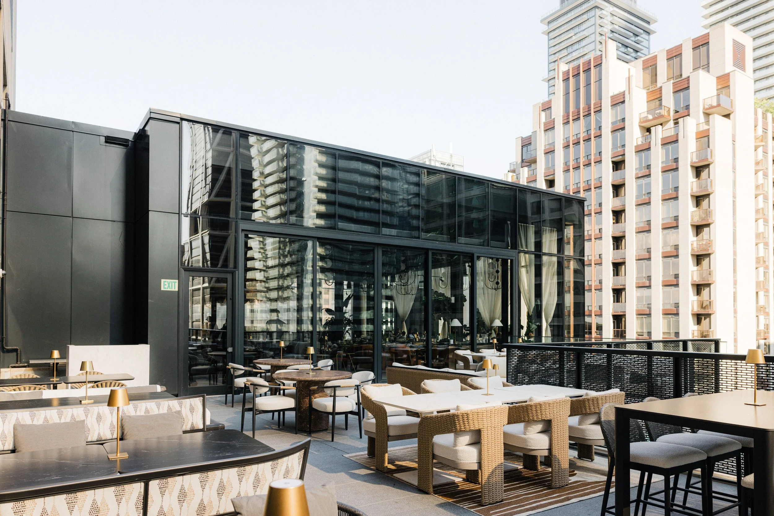 Outdoor rooftop dining area with tables, chairs, and small lamps, overlooking city buildings, including a modern glass restaurant and high-rise apartment buildings.