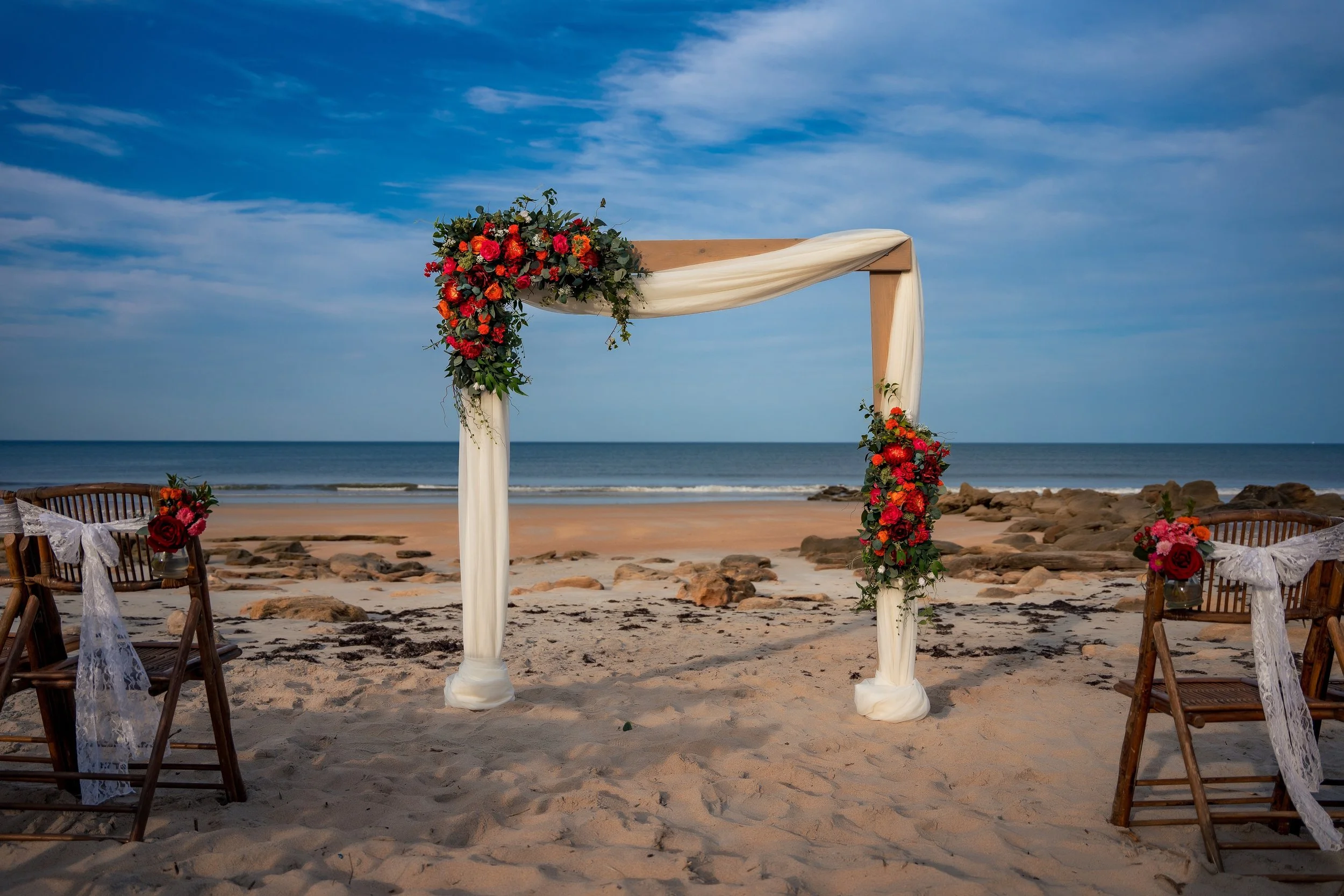 Beach wedding arch with floral decorations on sand, ocean and sky in background
