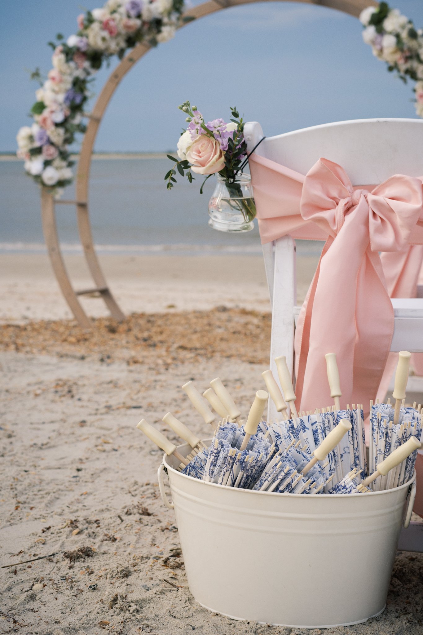 A white bucket filled with paper umbrellas, placed on sandy ground near a beach with a decorative arch floral arrangement in the background.