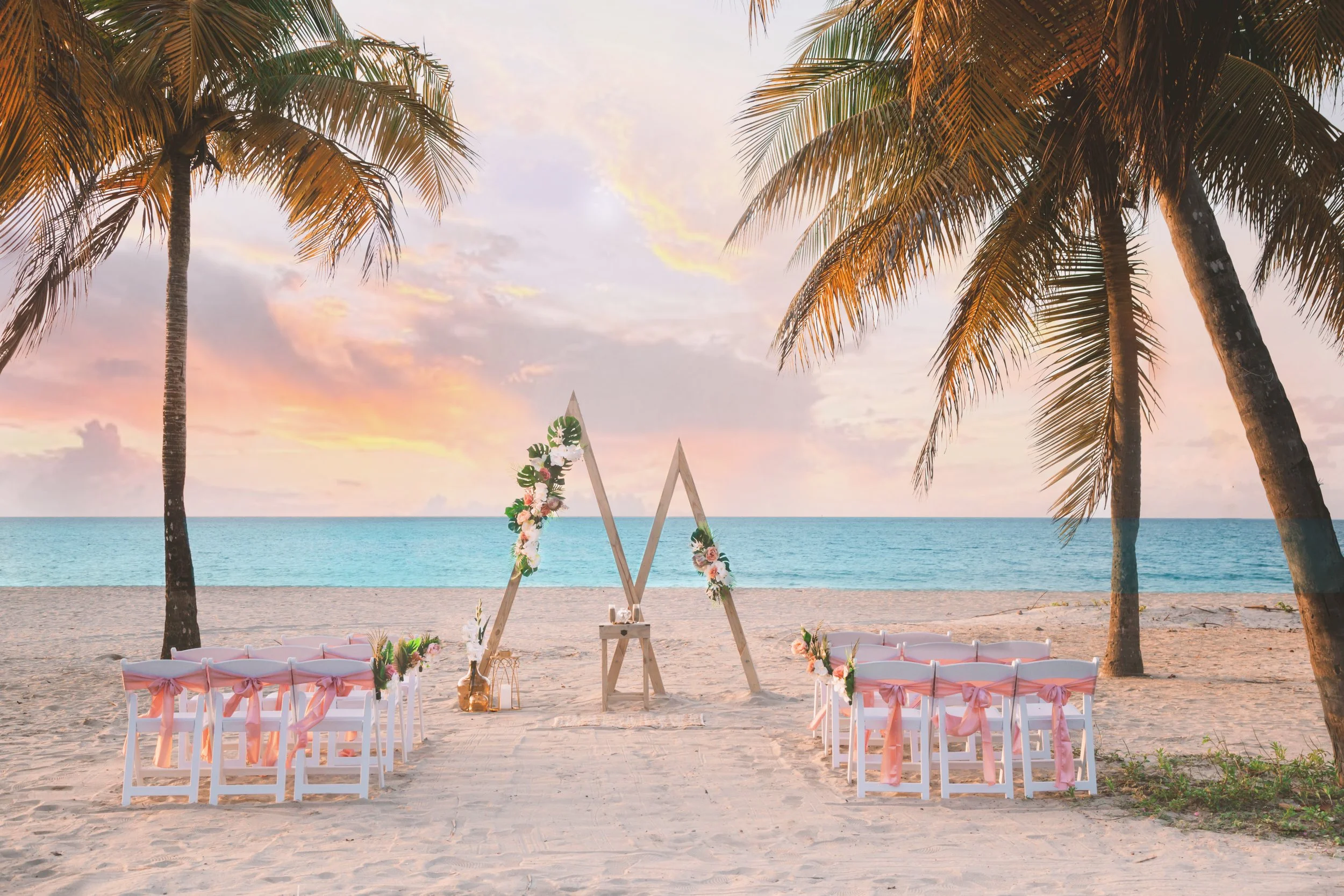 A wedding setup on a beach at sunset with chairs decorated with pink ribbons, a floral arch, and palm trees.