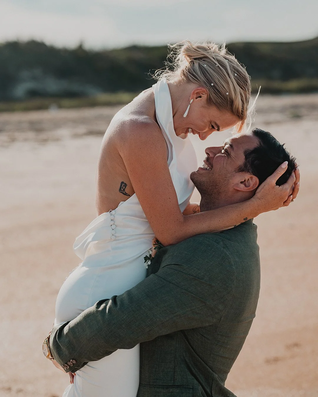 A man in a dark suit holding a woman in a white dress on a beach, both smiling and looking happy.
