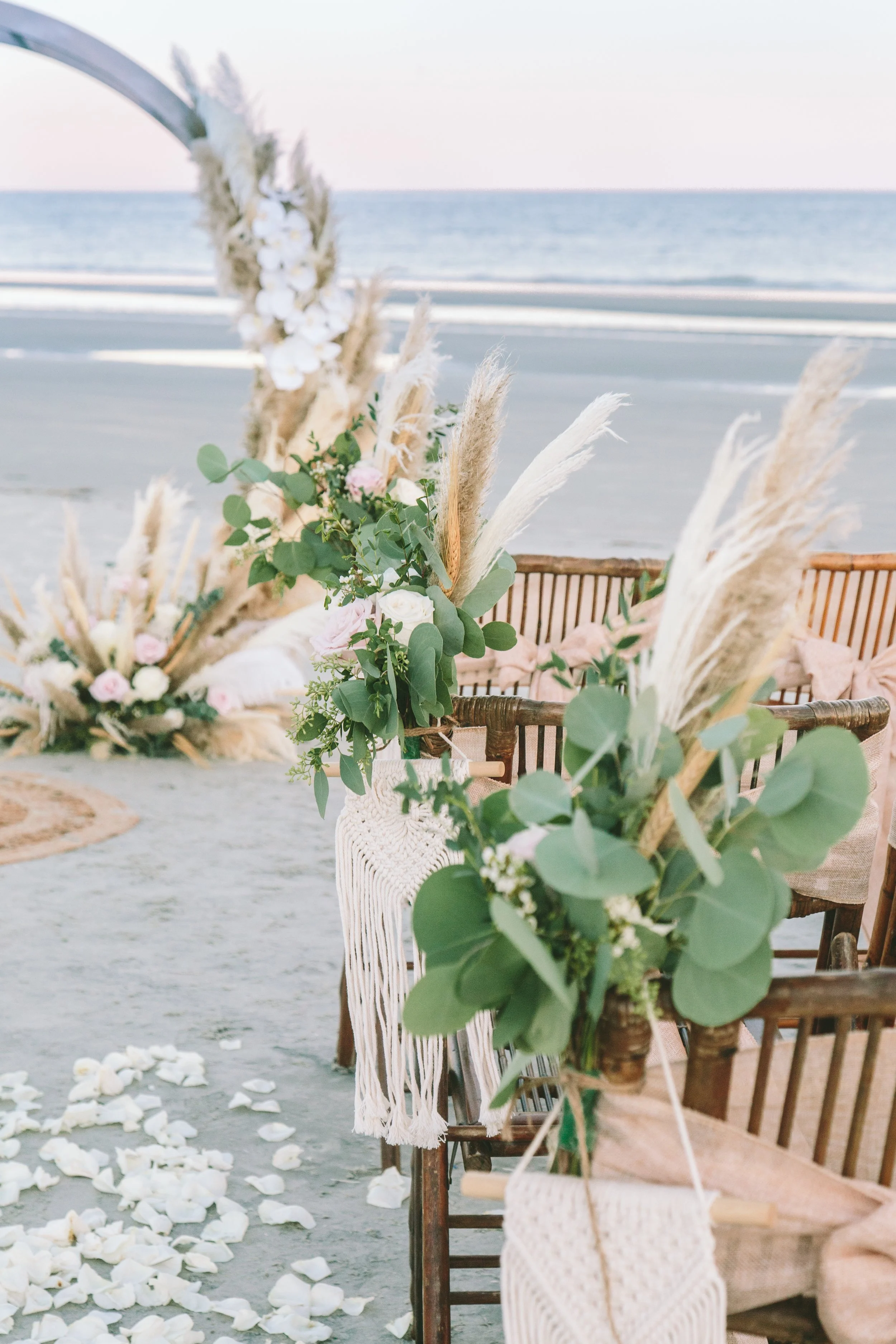 Beach wedding setup with floral decorations, chairs, and rose petals on sand, overlooking the ocean.