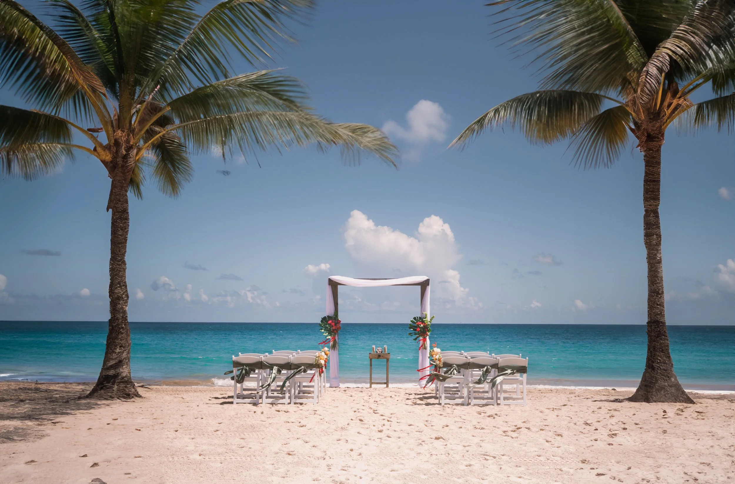 Beach wedding setup with white chairs, floral decorations, and a canopy, positioned between two palm trees overlooking the ocean.
