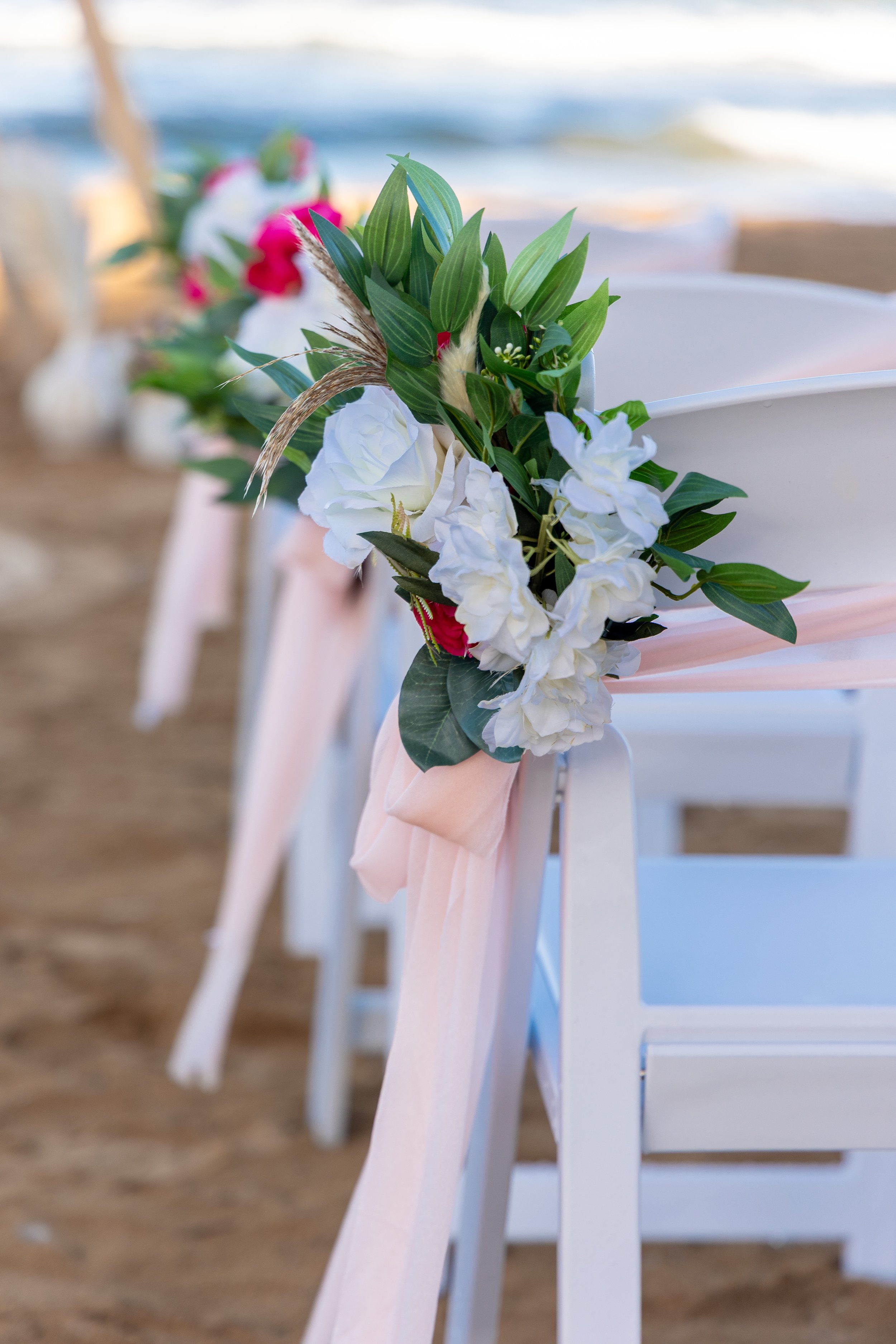 White chairs decorated with pink and white floral arrangements attached with pink ribbons, set up on a beach with sand and ocean waves in the background.