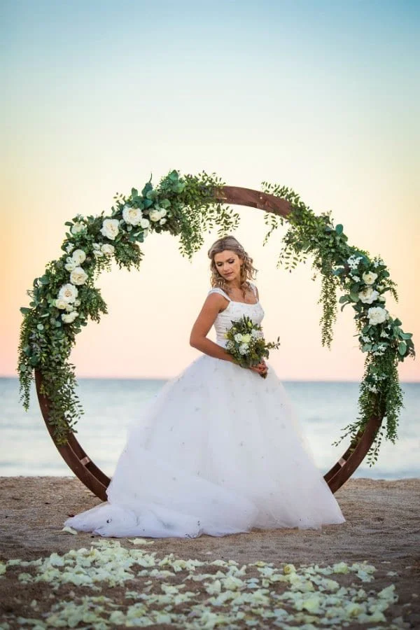 Bridal portrait of a woman in a white wedding dress holding a bouquet, standing in front of a large floral arch on a beach during sunset.