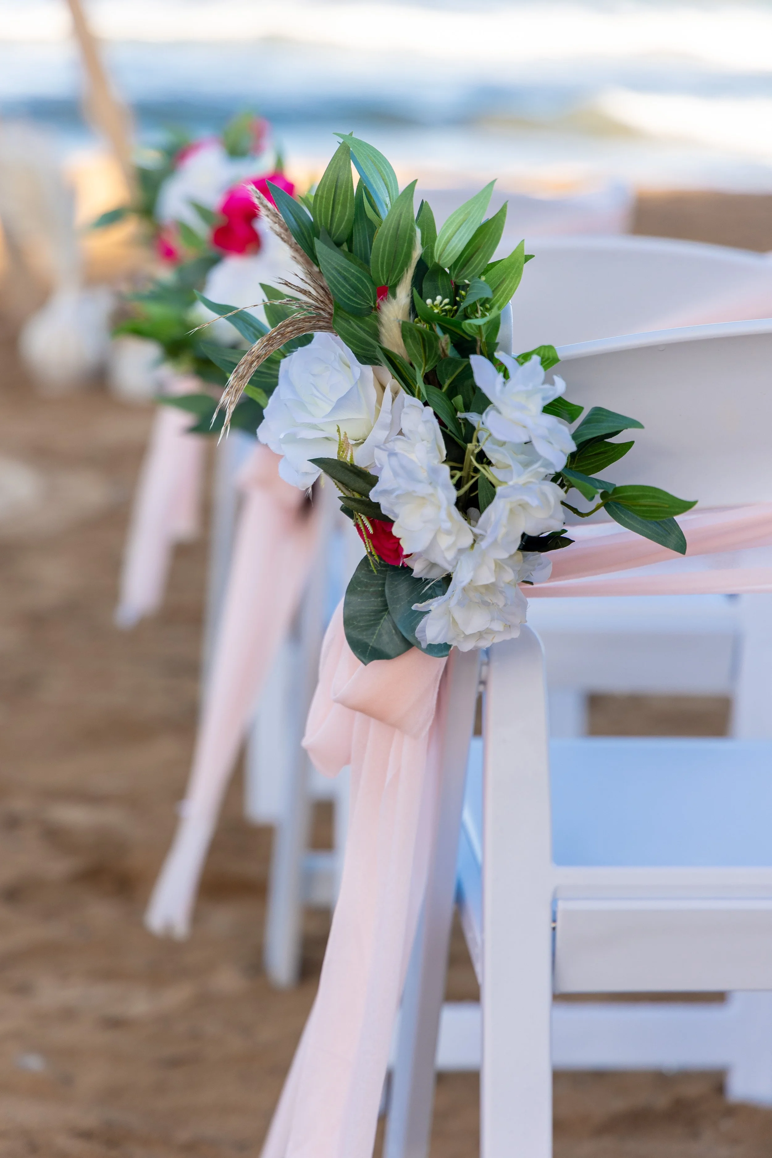 White chairs decorated with flowers and ribbons, lined up on a sandy beach for a wedding ceremony, near the ocean with waves in the background.