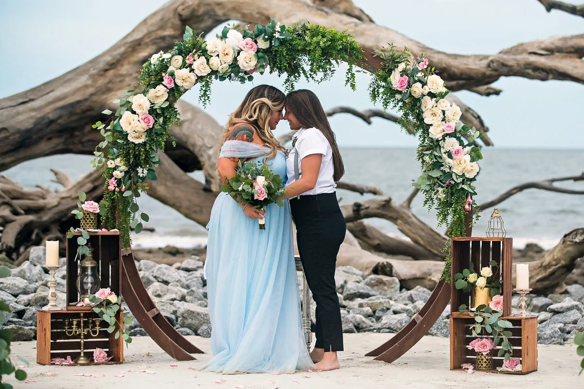Two women standing close together on a beach, under a floral arch, holding a bouquet of flowers, with driftwood and rocks in the background.