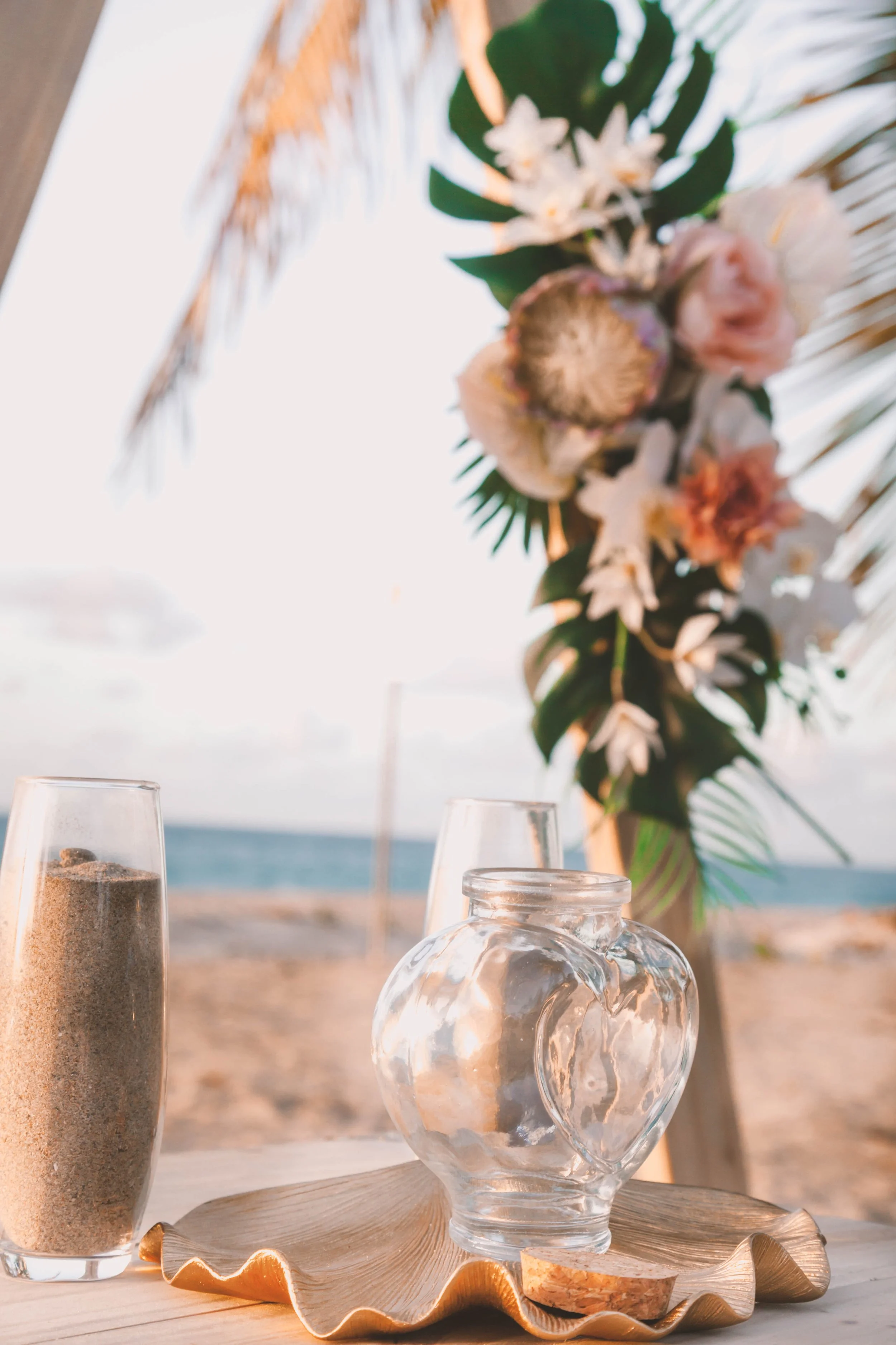 Beachside table with glass vase, filled with sand, and empty glass cups, decoratively arranged on a shell-shaped tray, with a bouquet of tropical flowers and green leaves in the background, and a blurred ocean view.