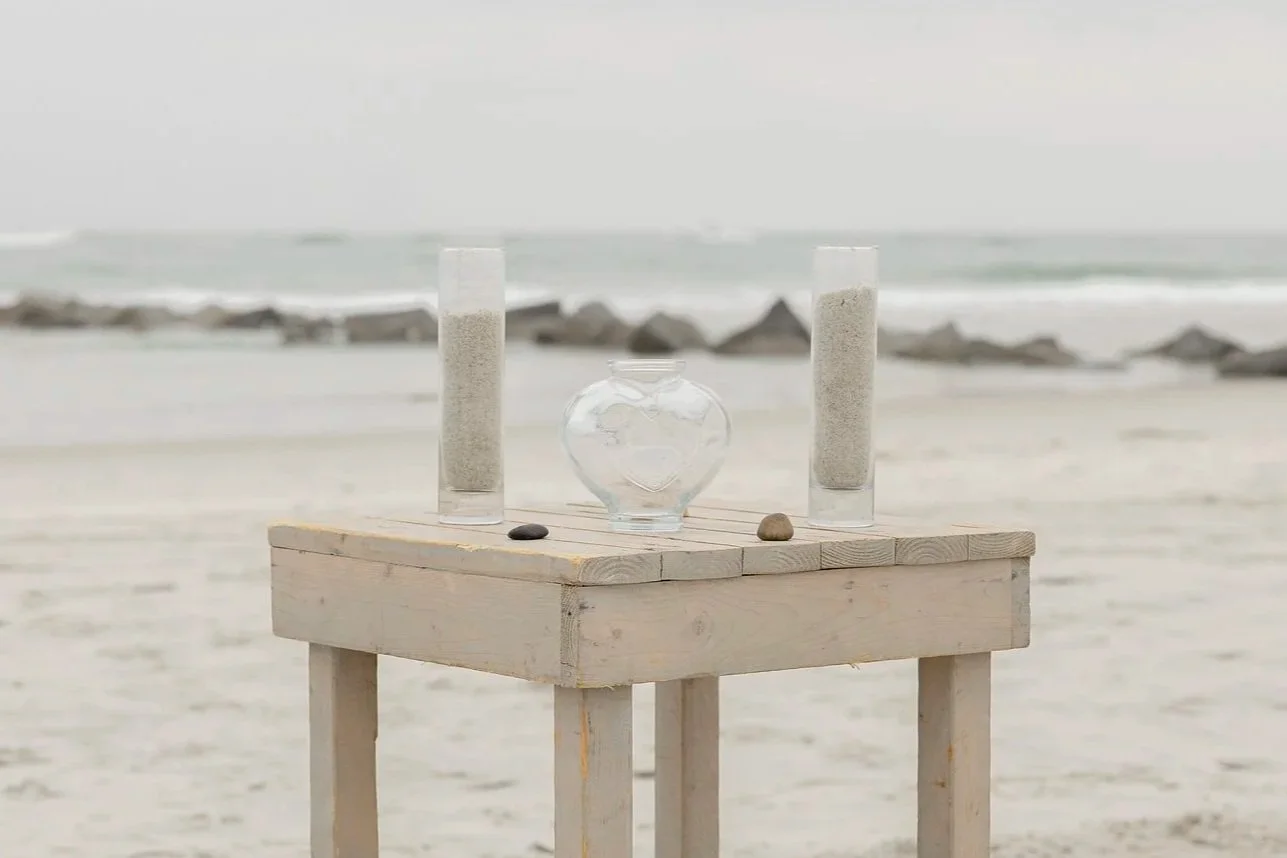 A small wooden table on a beach with a glass heart-shaped vase, two tall glass cylinders filled with sand, and three small stones, with the ocean and rocks in the background.