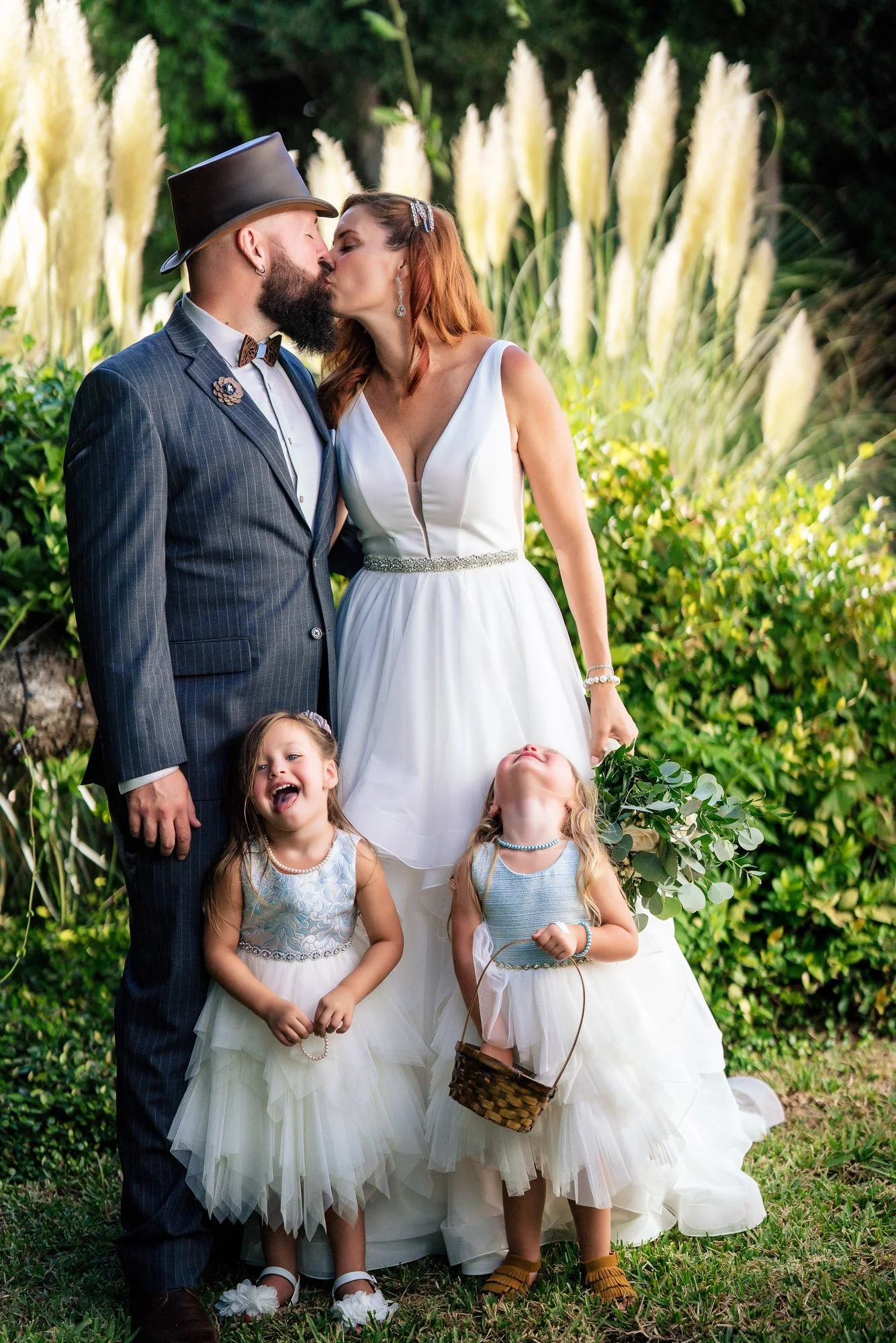 A couple dressed in wedding attire kiss outdoors, surrounded by greenery, with three young girls in white dresses standing in front of them.