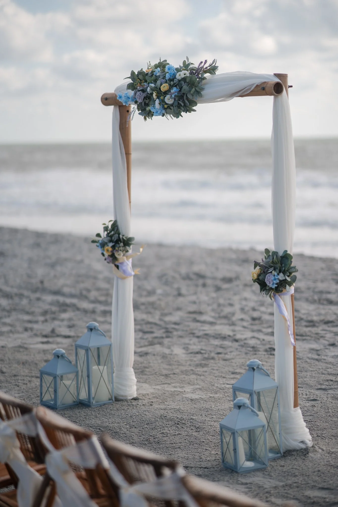 Beach wedding arch decorated with white fabric and floral arrangements with blue, white, yellow, and purple flowers, surrounded by glass lanterns with candles at sunset.