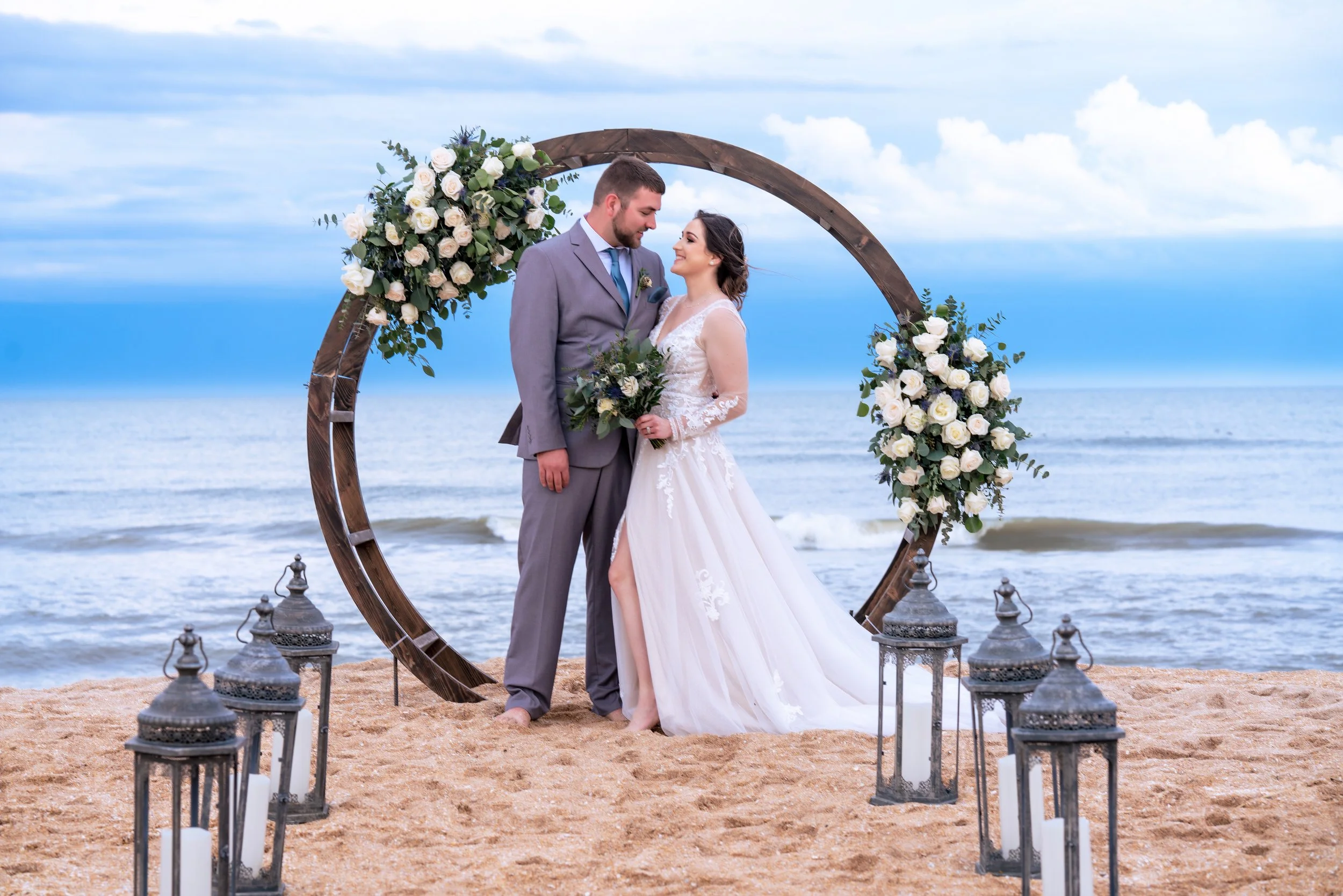 A bride and groom standing on the beach with a floral wedding arch behind them, decorated with white roses and greenery, surrounded by lanterns with candles, during a seaside wedding.