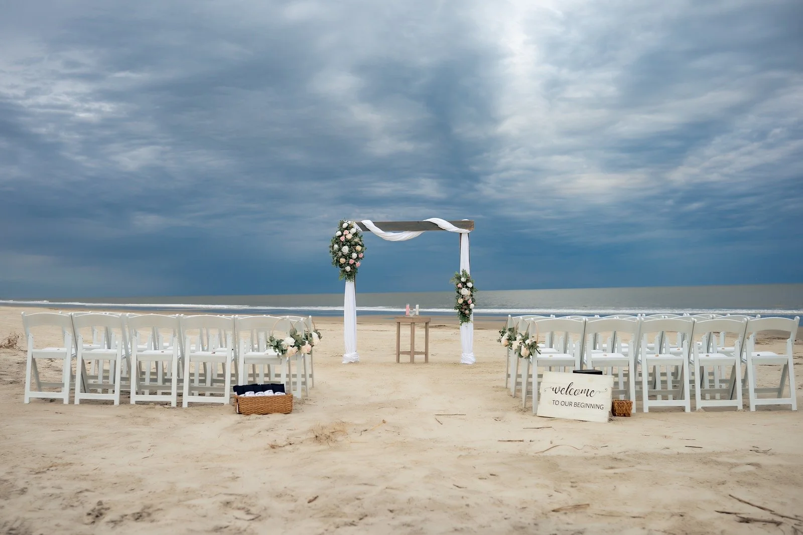 Beach wedding setup with white chairs, floral decorations, and a wooden arch under a cloudy sky, facing the ocean.