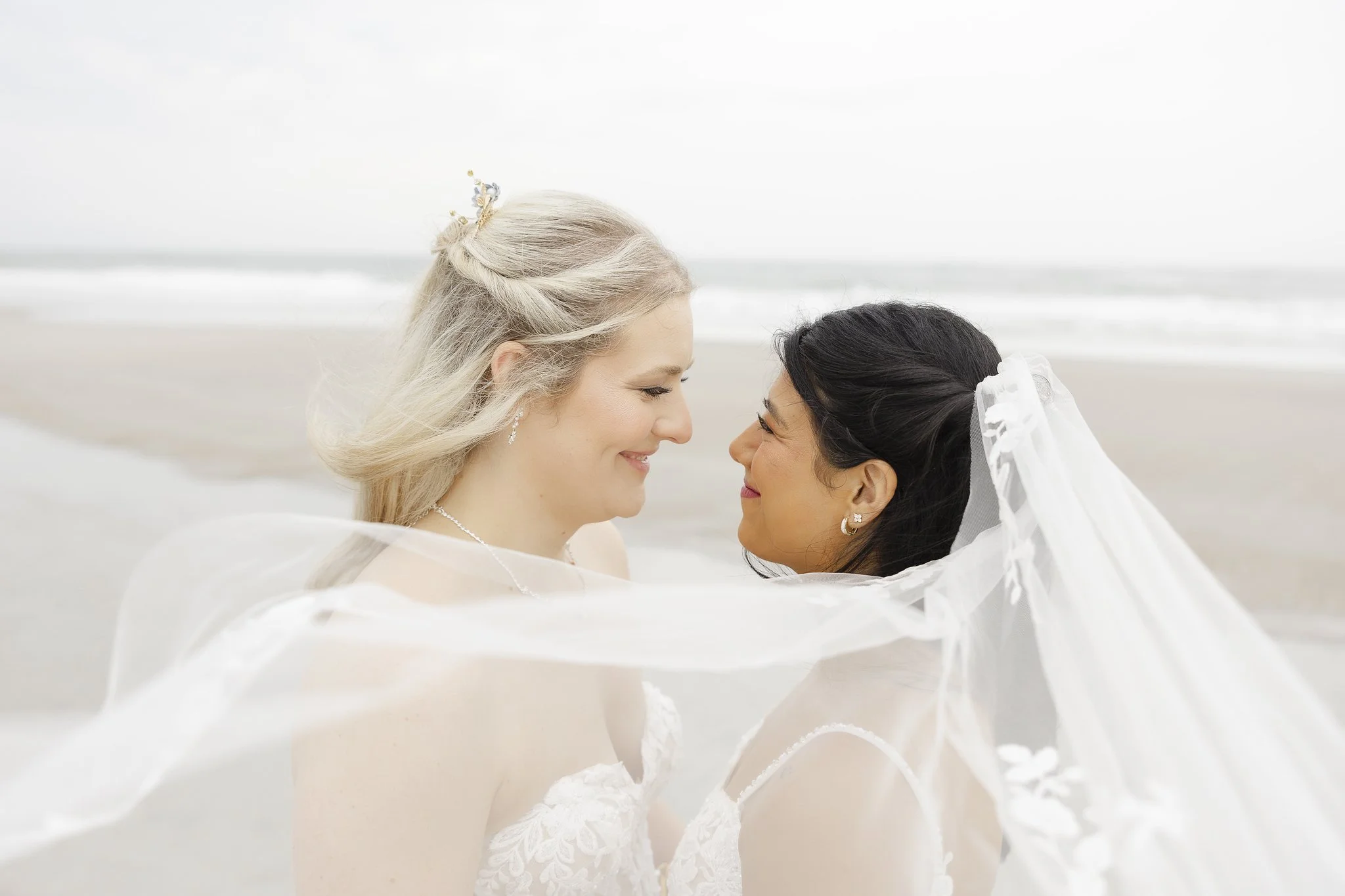 Two women with wedding dresses touching foreheads on a beach, with waves in the background.