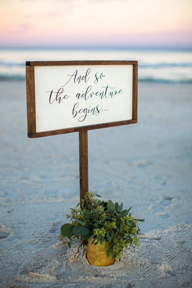 A wooden sign on a beach reads, 'And so the adventure begins...' in elegant script, surrounded by a small green plant in a yellow pot, with ocean waves and a pink sky in the background.