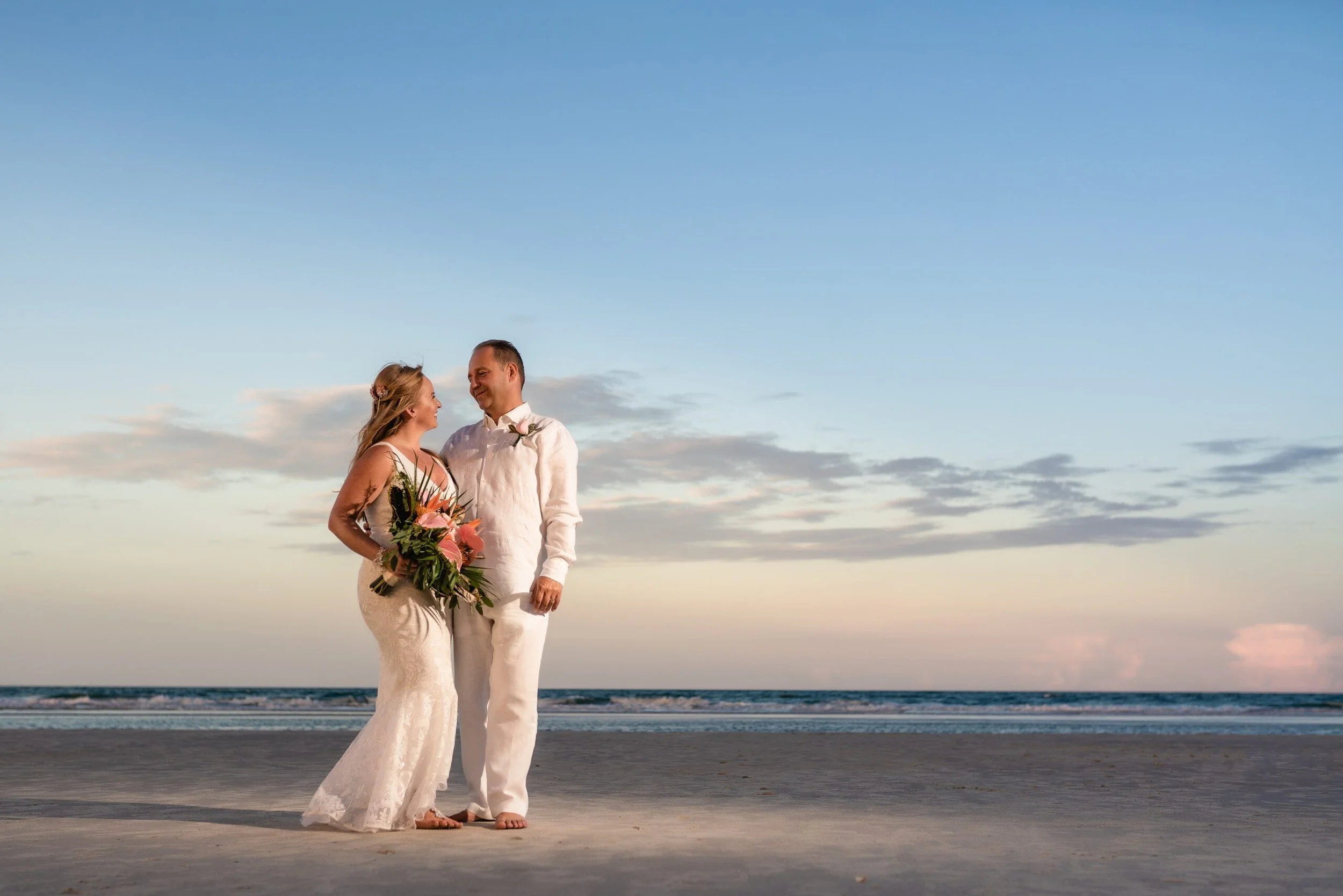 A bride and groom stand on a beach at sunset, gazing at each other. The bride wears a lace wedding dress and holds a bouquet of pink flowers, while the groom wears a white shirt and beige pants. The sky is partly cloudy with soft light illuminating the scene.