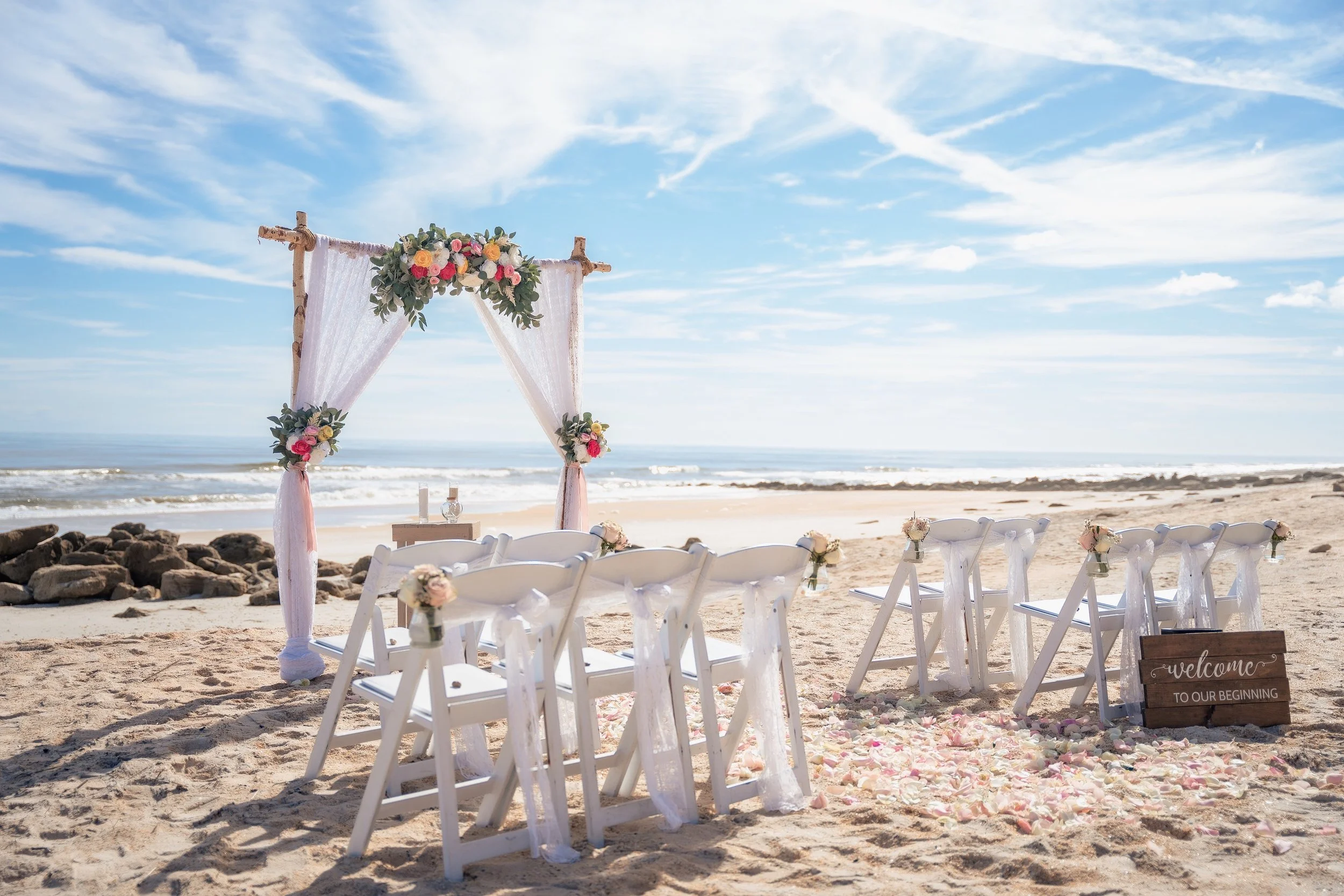 Beach wedding setup with a floral arch, white chairs decorated with flowers, and a sign that says "Welcome to our beginning" on sandy beach with blue sky and ocean in the background.